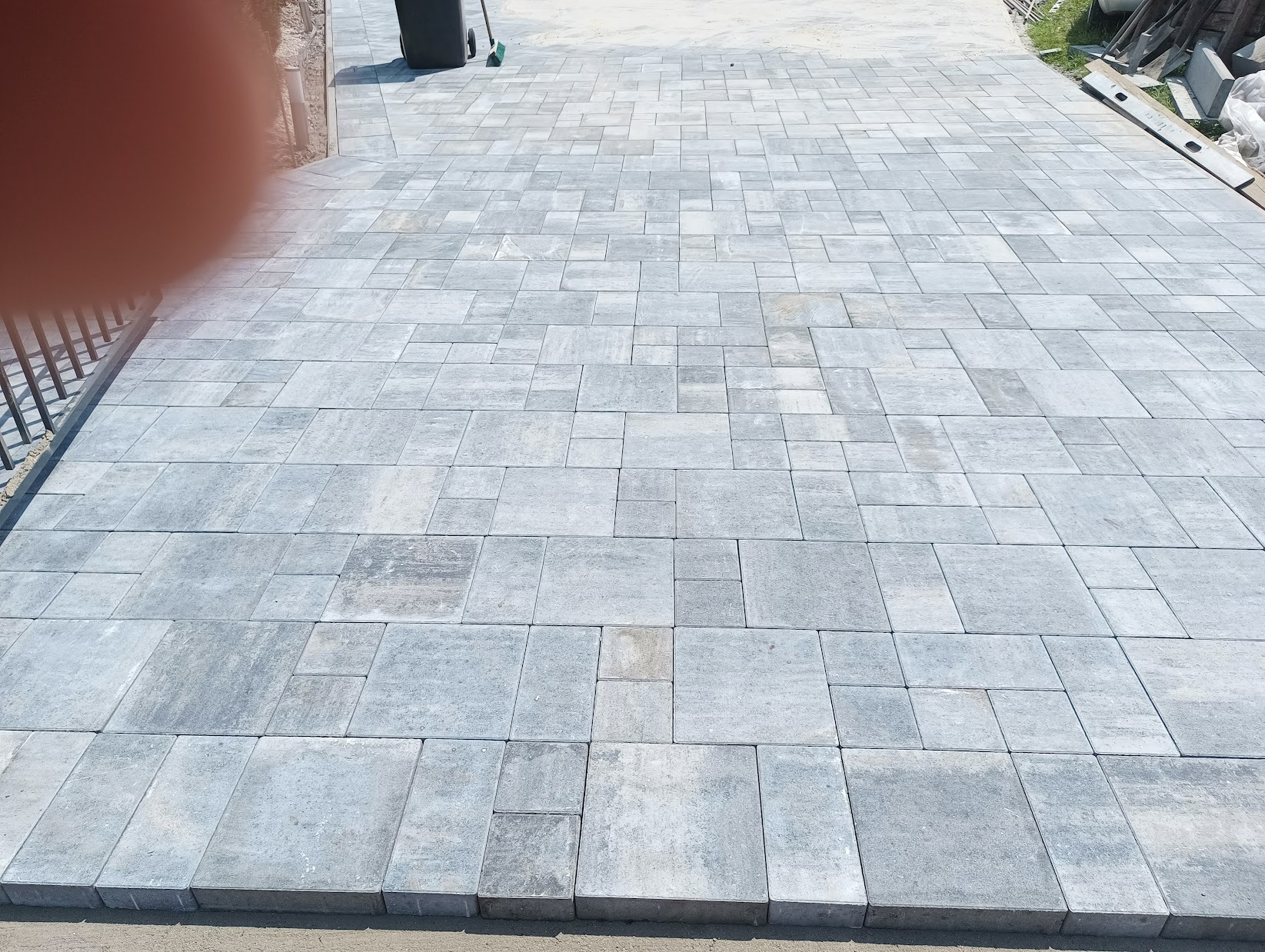 Świeżo ułożona, szara kostka brukowa w różnych rozmiarach, tworząca wzór na posesji, z widocznym fragmentem ogrodzenia i elementami krajobrazu w tle.