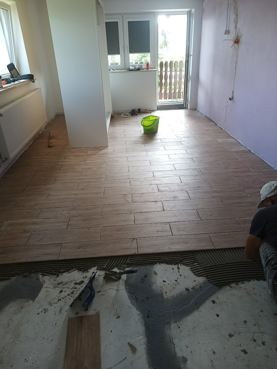 Układanie podłogi z płytek imitujących drewno w jasnym pomieszczeniu, widoczne narzędzia glazurnicze i częściowo ułożona podłoga, w tle widok na balkon.