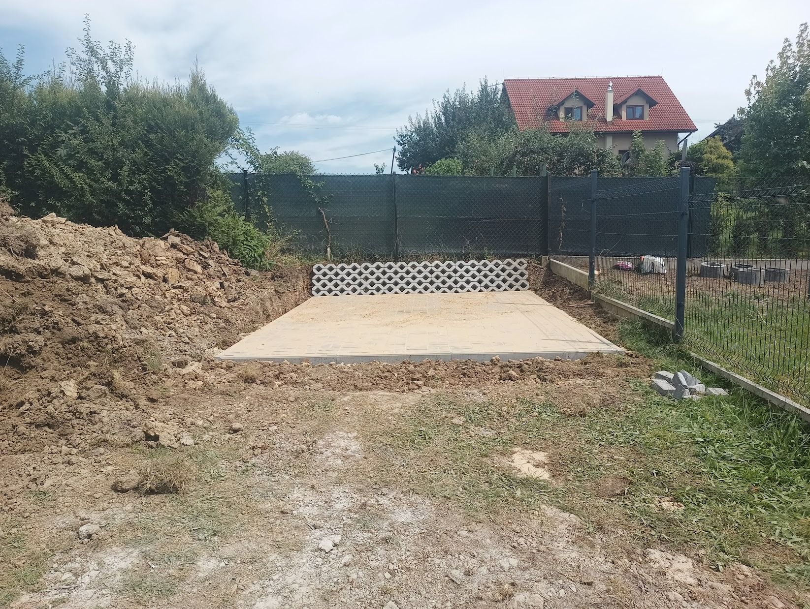 Przygotowany podkład z obrzeżami pod taras lub kostkę brukową, widoczny stos ziemi, ogrodzenie z siatki i dom w tle.
