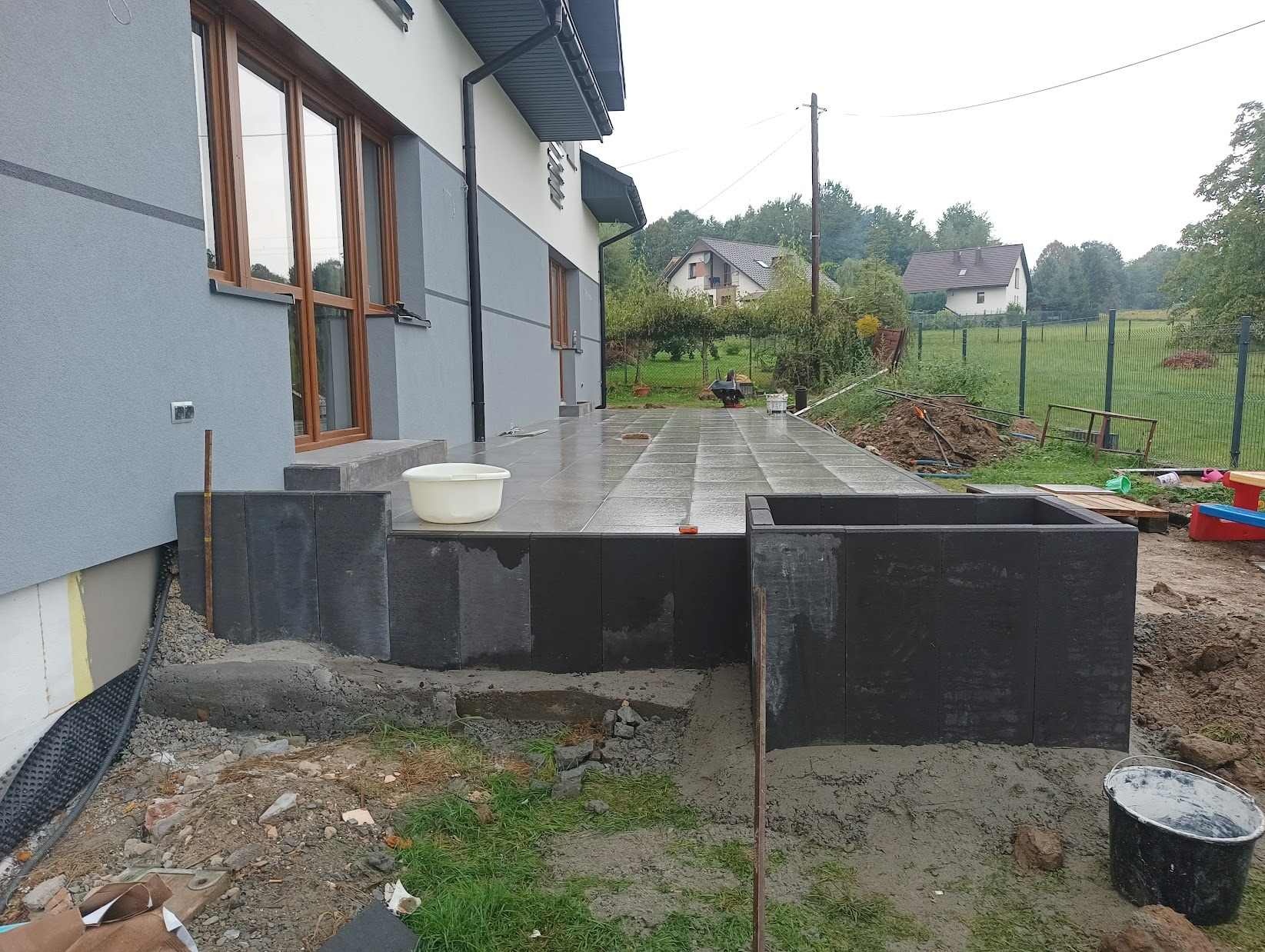 Budowa tarasu z szarych płyt na zewnątrz domu, widoczne bloczki betonowe, biała miska i wiadro, teren wokół w trakcie prac ziemnych.