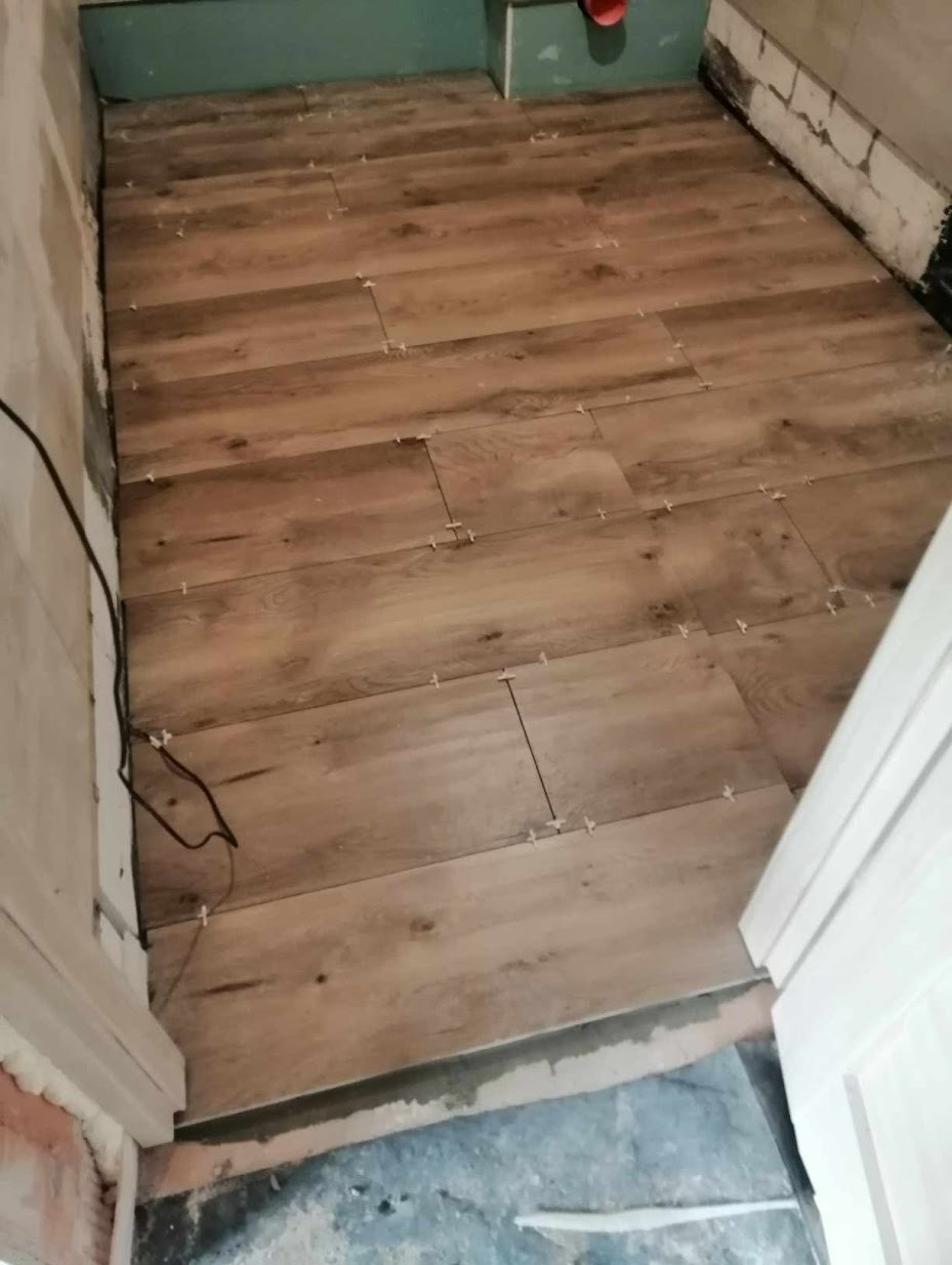 Układanie podłogi z płytek imitujących drewno w trakcie prac wykończeniowych, widoczne krzyżyki dystansowe i przewód elektryczny przy ścianie.