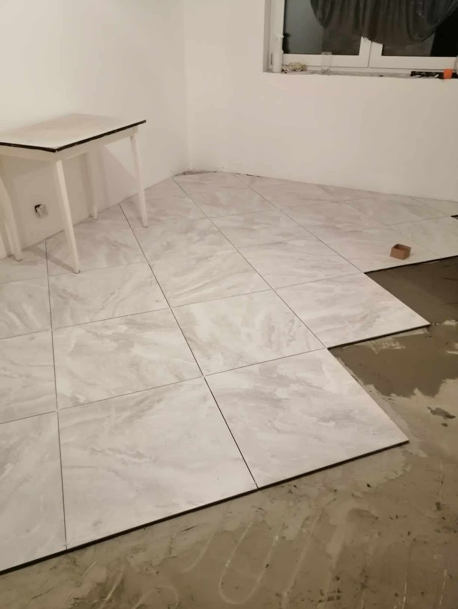 Wnętrze pokoju w trakcie układania jasnych, marmurkowych płytek podłogowych; widoczny fragment podłogi z nieułożonymi płytkami i śladami kleju oraz biały stolik pomocniczy w rogu pomieszczenia.
