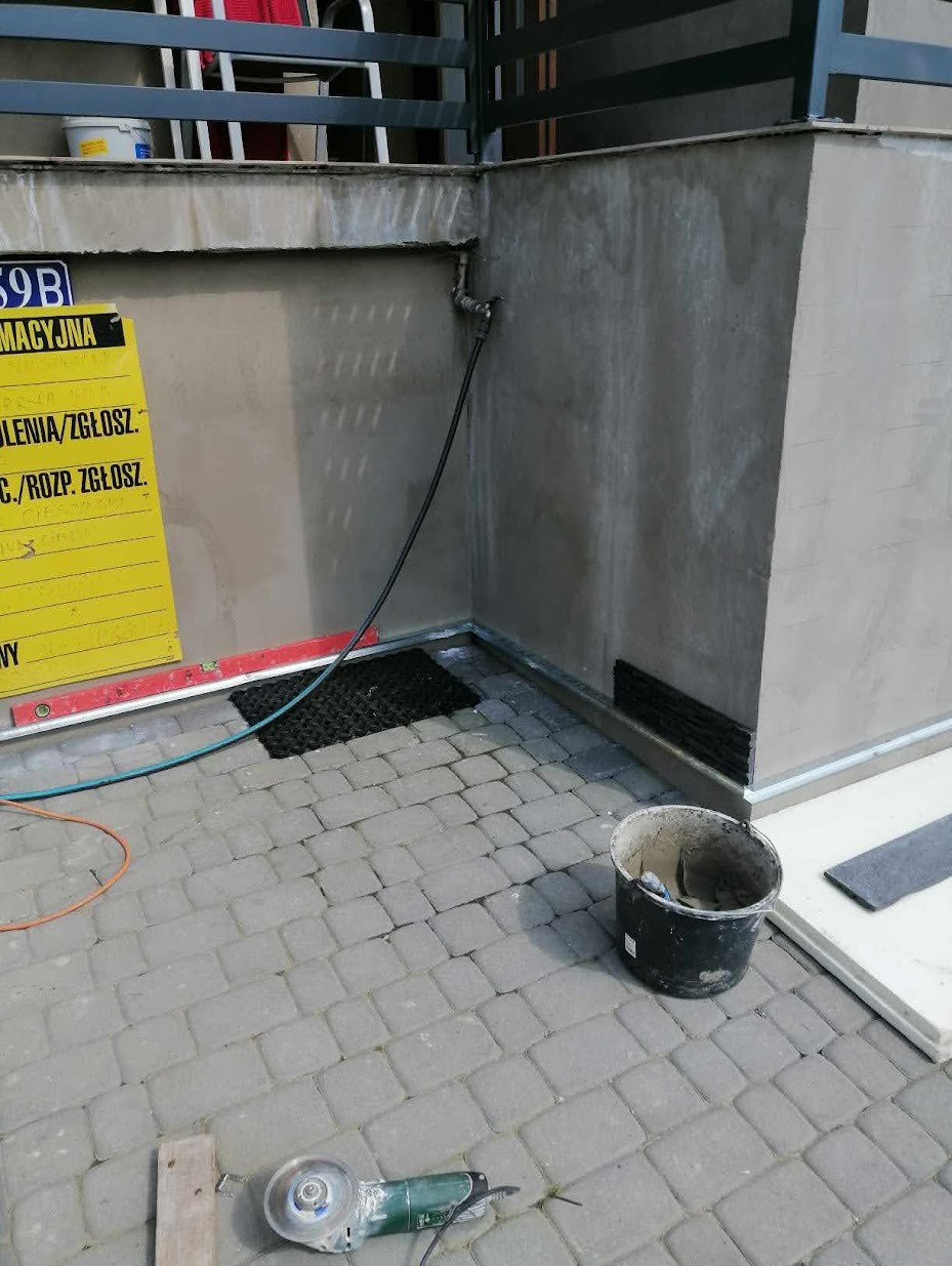 Szara kostka brukowa ułożona przy ścianie budynku z widoczną rurą, żółtą tablicą informacyjną, poziomica, wiadro z zaprawą i szlifierką kątową na pierwszym planie.