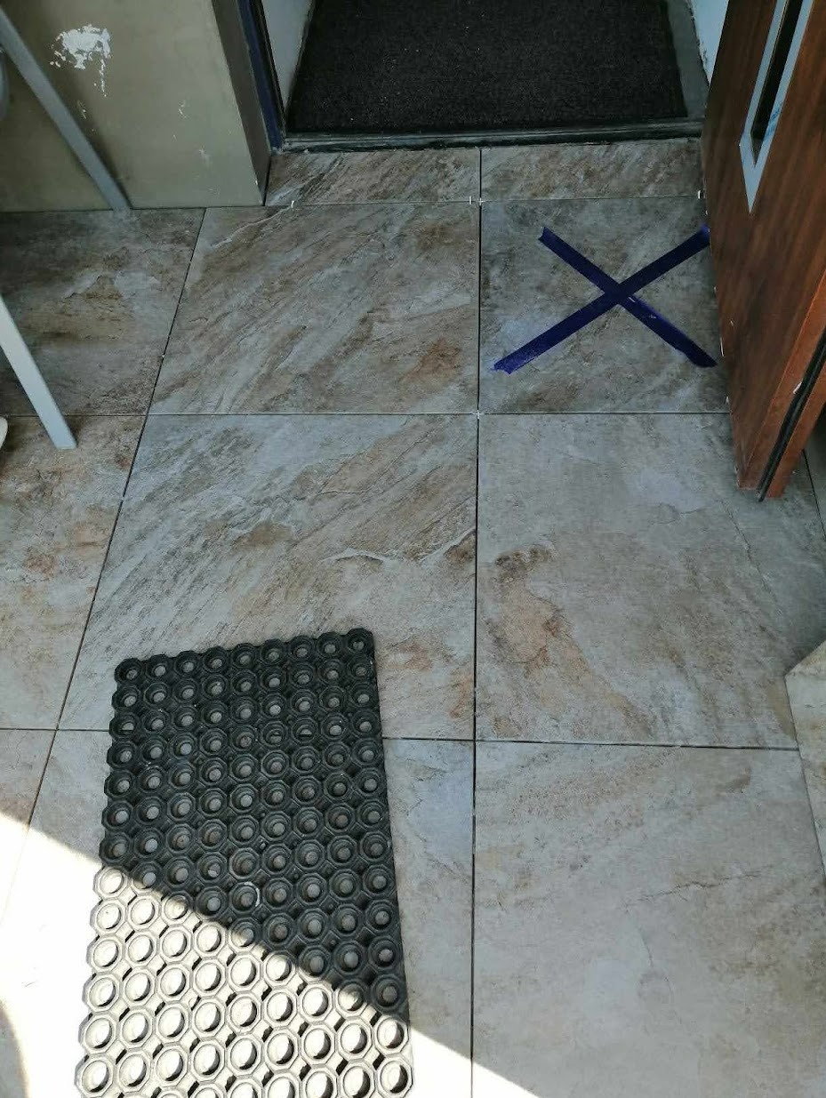 Ułożone płytki podłogowe imitujące marmur, z niebieską taśmą w kształcie X na jednej z płytek i gumową wycieraczką w rogu, przy wejściu.