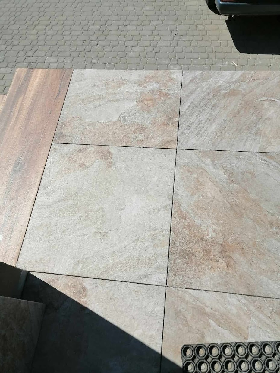 Ułożone kwadratowe płyty tarasowe w odcieniach beżu i szarości, z widoczną strukturą kamienia, sąsiadujące z drewnopodobną płytą i szarą kostką brukową. W rogu widoczna wycieraczka.