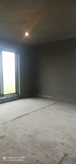 Surowe wnętrze pokoju po nałożeniu szarego tynku na ściany, widoczne niedoskonałości powierzchni i surowy beton na podłodze, pojedyncza żarówka zwisająca z sufitu.