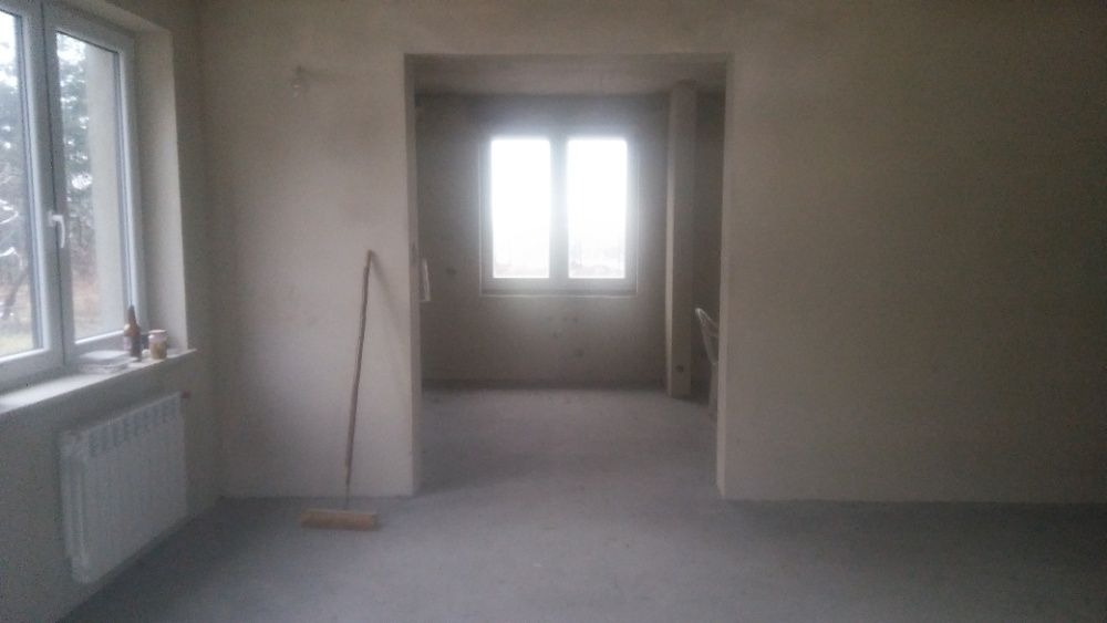 Wnętrze surowego pomieszczenia po tynkowaniu maszynowym, widoczne ślady świeżego tynku na ścianach, grzejnik pod oknem, oraz szczotka oparta o ścianę.