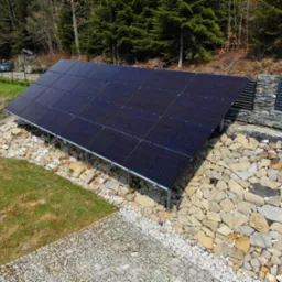 Instalacja fotowoltaiczna na skalnym podłożu, widok z góry, w tle las iglasty i samochód za ogrodzeniem.