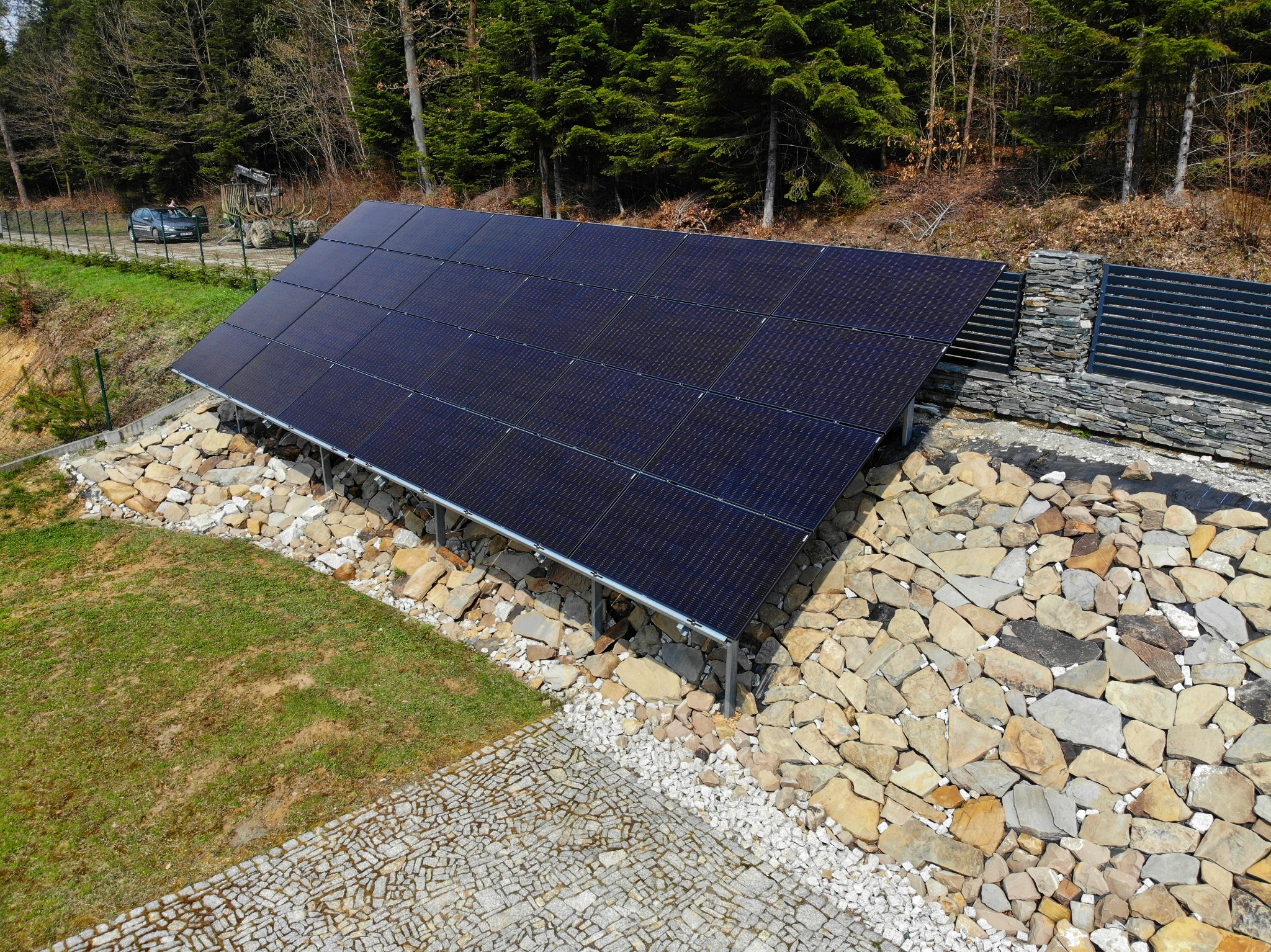 Instalacja fotowoltaiczna na skalnym podłożu, widok z góry, w tle las iglasty i samochód za ogrodzeniem.
