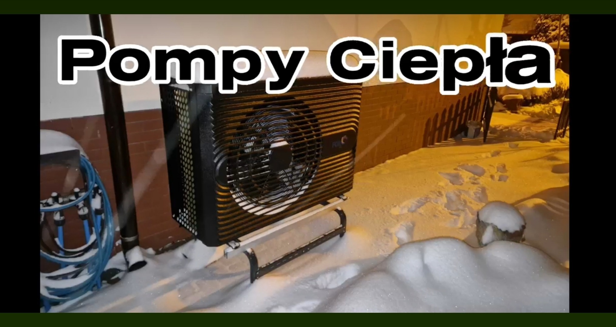 Zewnętrzna jednostka pompy ciepła stojąca na śniegu w zimowej scenerii, z widoczną elewacją budynku w tle i oświetleniem ulicznym.