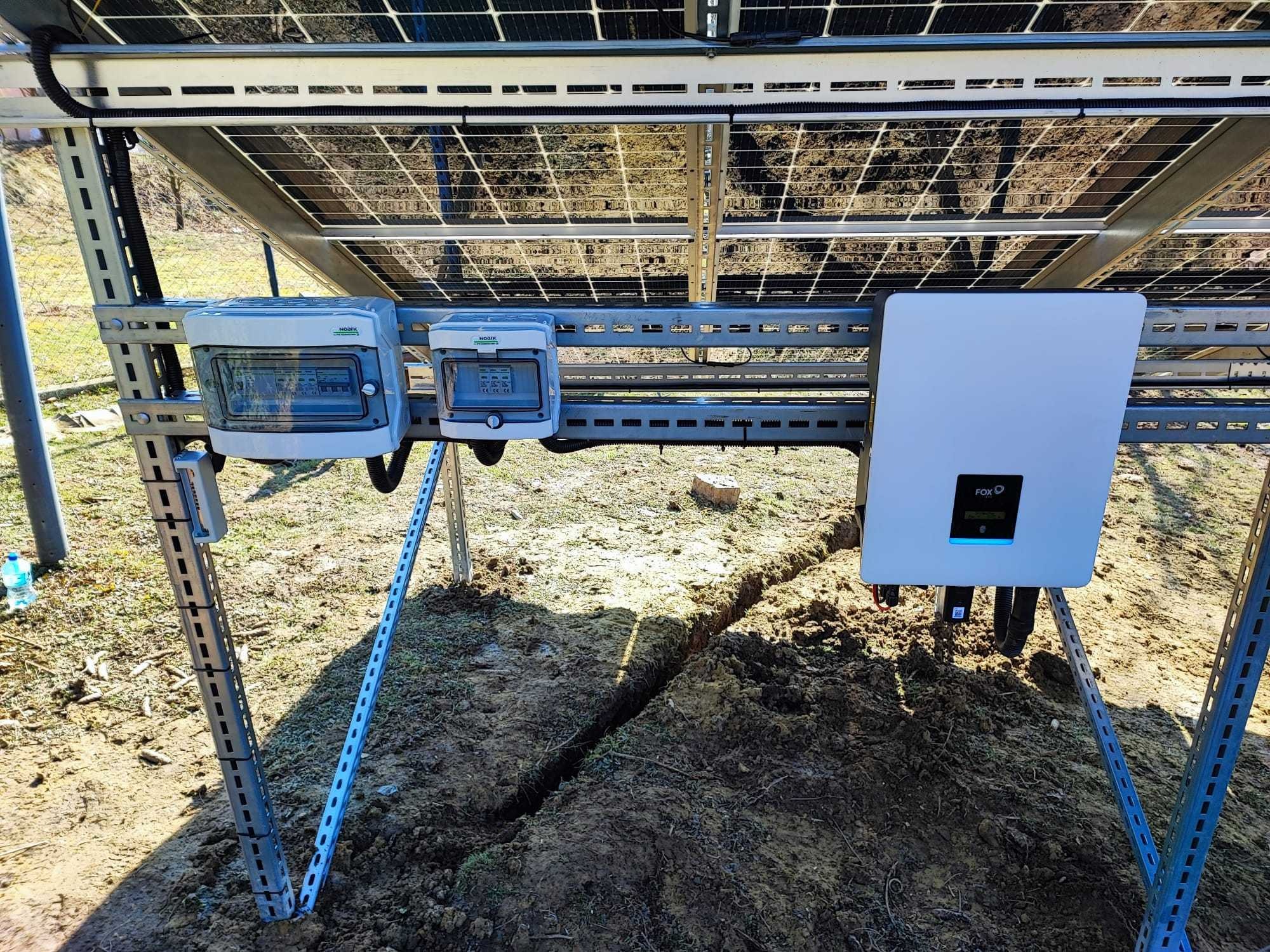 Montaż instalacji fotowoltaicznej: widok od spodu na panele, metalową konstrukcję wsporczą, dwa białe rozdzielnice elektryczne i biały falownik Fox ESS, z wykopanym rowem w ziemi.