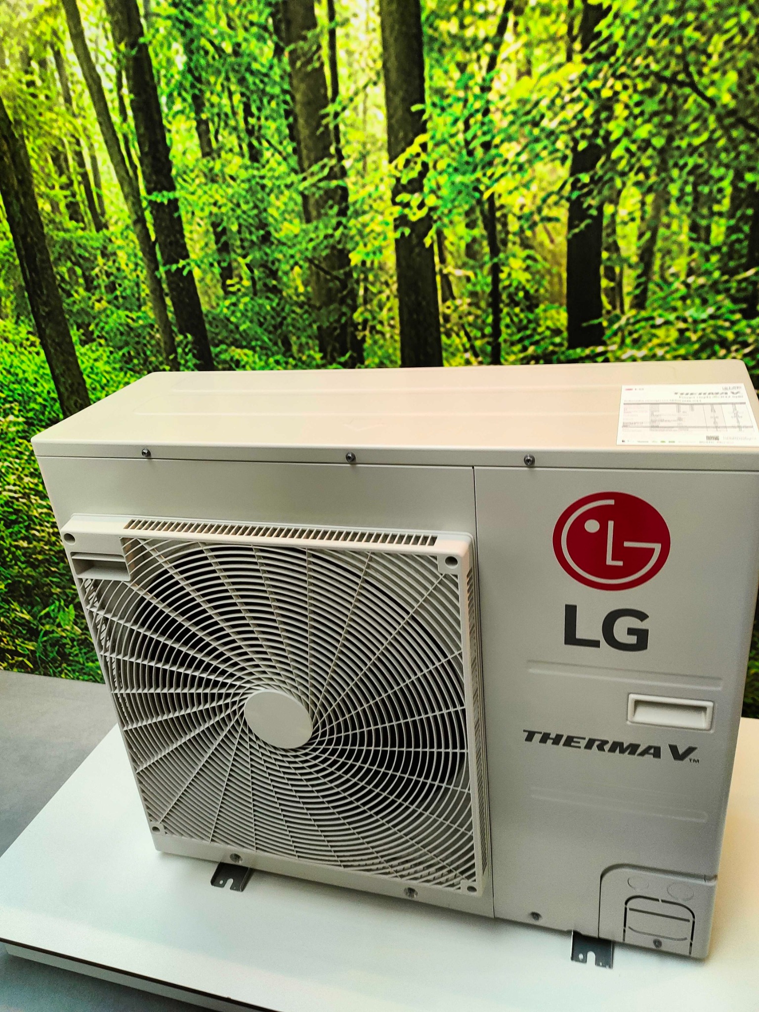 Kompaktowa jednostka zewnętrzna pompy ciepła LG Therma V, z wyraźnym logo, umieszczona na białym podłożu na tle fototapety imitującej las.