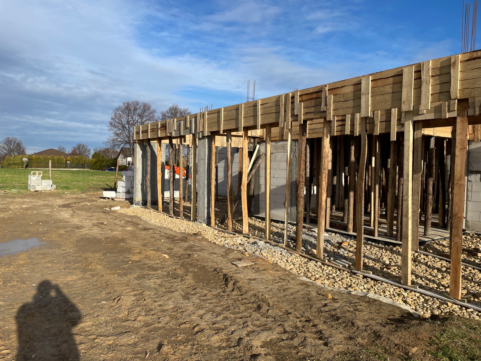 Wznoszenie ścian domu z betonu komórkowego, widoczne szalunki i zbrojenie, teren budowy z gruzem i śladami opon, cień fotografa w lewym dolnym rogu.