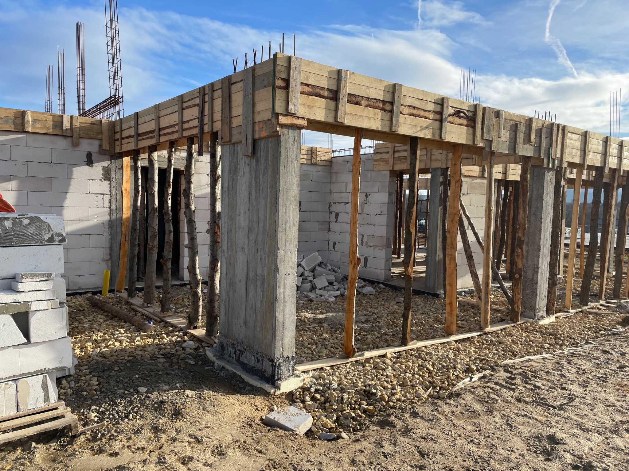 Surowy stan budowy domu z betonu komórkowego, widoczne szalunki i zbrojenia pionowe, teren wokół wysypany żwirem, niebo z lekkimi chmurami w tle.