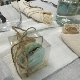 Ustawienie stołu z dekoracyjnymi serwetkami z koronką, szklanką, dzbankiem z wodą i pudełeczkiem z makaronikami związanym sznurkiem.