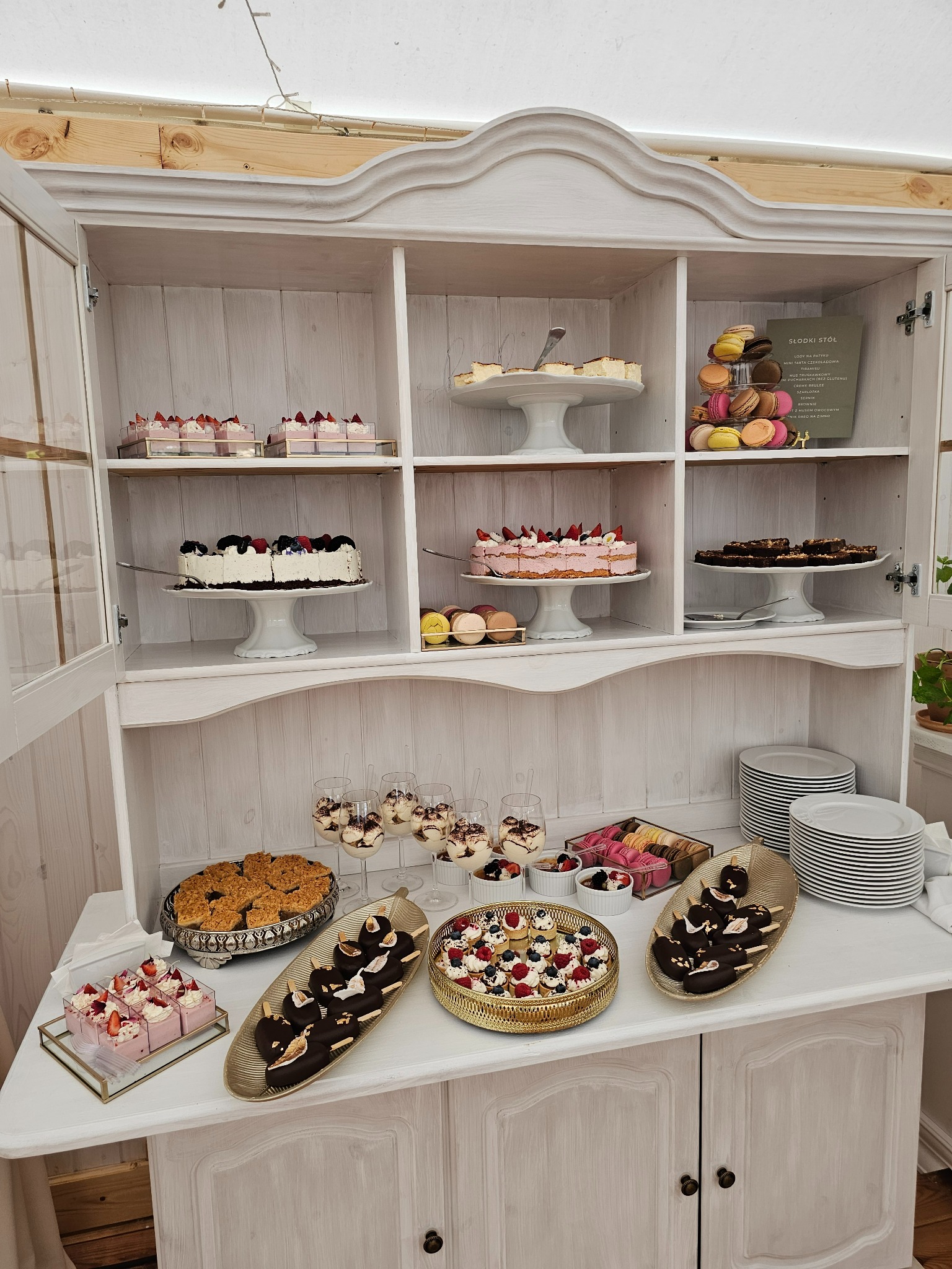 Słodki stół w Krakowie, z tortami, deserami w szklankach, makaronikami i ciastkami, umieszczony w białym kredensie w stylu vintage, z widocznym menu słodkiego stołu.