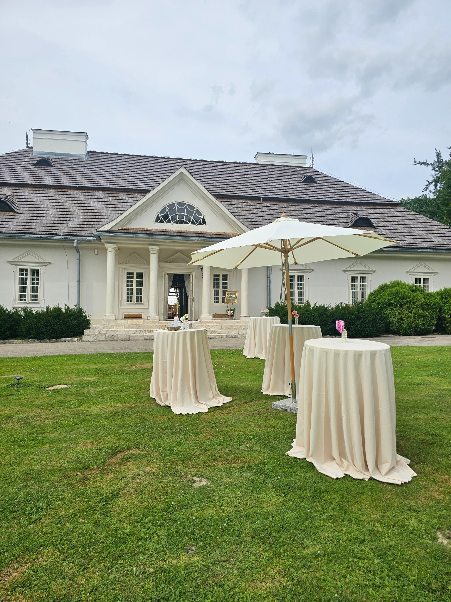 Cztery okrągłe stoliki koktajlowe z kremowymi obrusami, ustawione na trawniku przed klasycystycznym budynkiem z kolumnami i białymi ścianami, przygotowane na wydarzenie plenerowe.