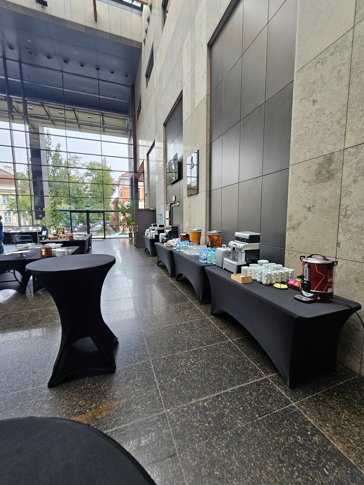 Sala konferencyjna z długimi stołami przykrytymi czarnymi obrusami, na których znajdują się dystrybutory z napojami, ekspresy do kawy, szklanki, filiżanki i drobne przekąski. W tle widoczne duże...