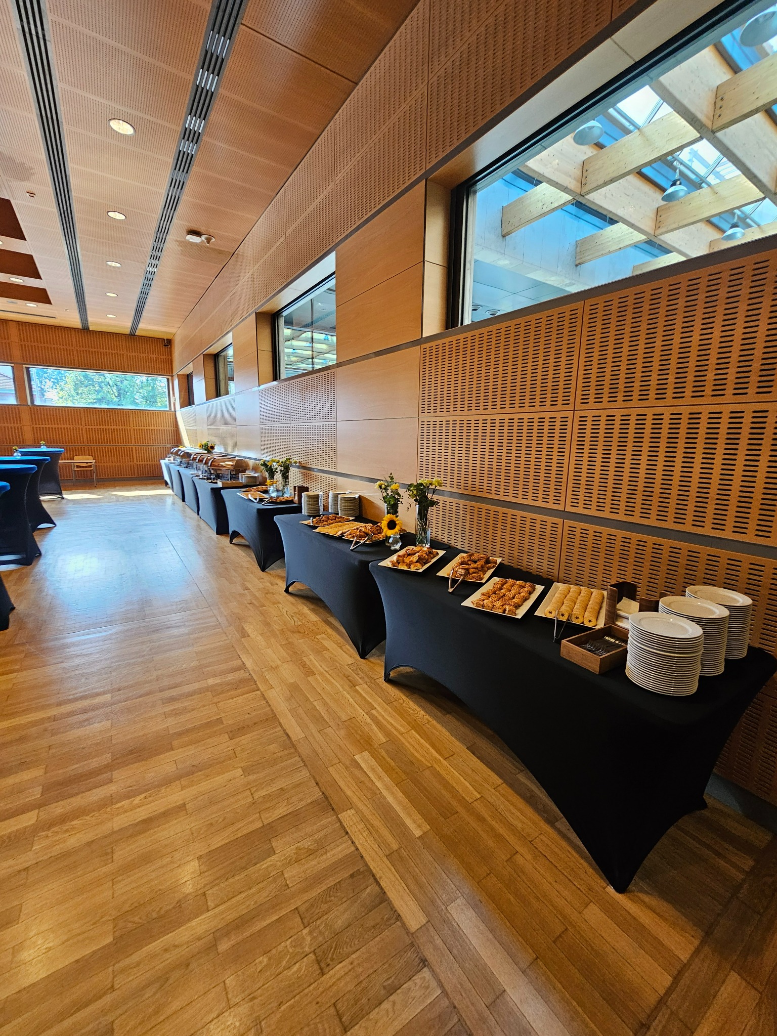 Długi stół bufetowy z czarnym obrusem, zastawiony różnorodnymi potrawami, talerzami i kwiatami w przestronnym wnętrzu z drewnianą boazerią i dużymi oknami.