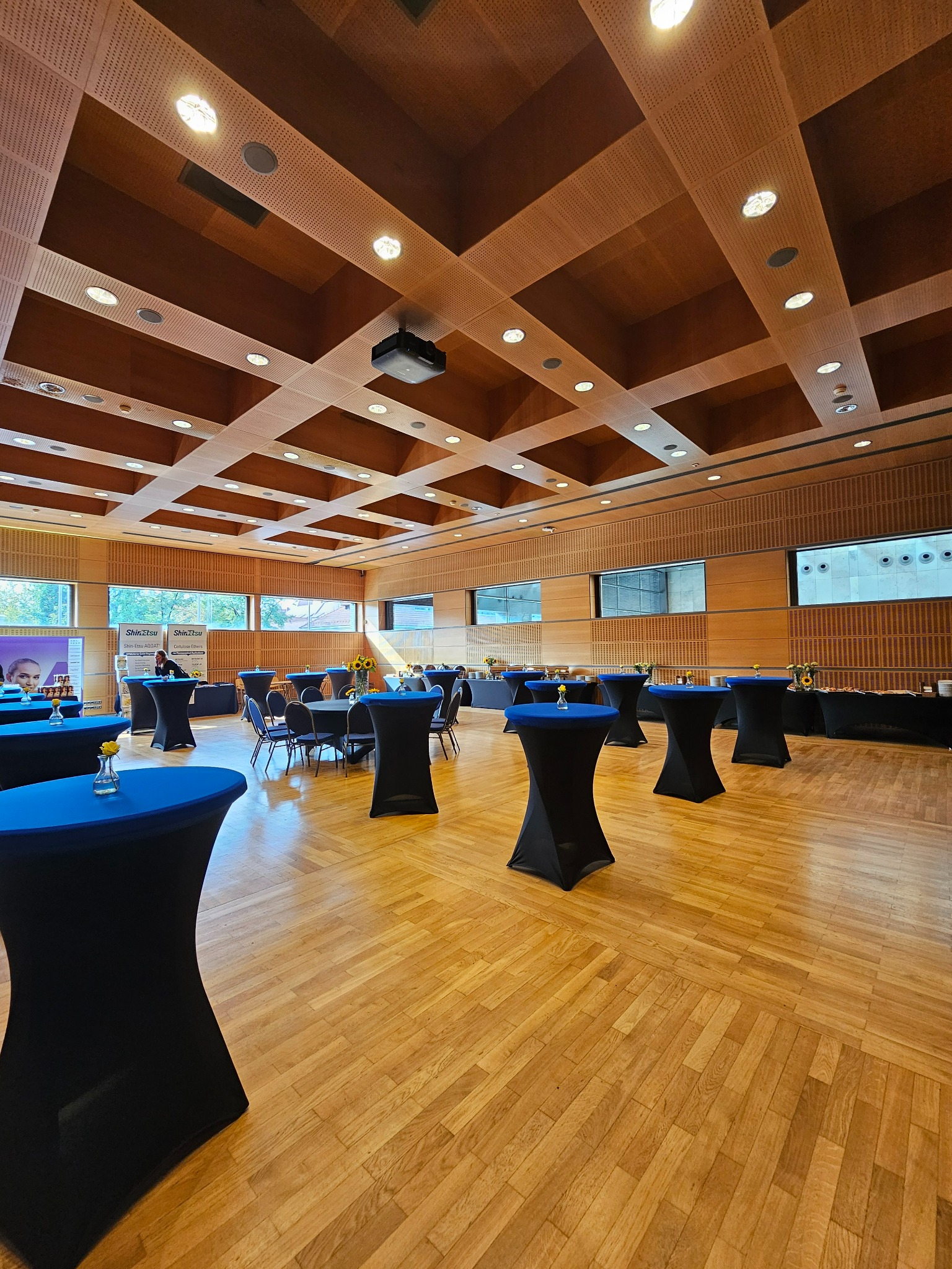Sala konferencyjna z drewnianym parkietem i sufitami kasetonowymi, przygotowana na wydarzenie z wysokimi stolikami w czarnych pokrowcach z niebieskimi blatami i kwiatami w wazonach, w tle catering...
