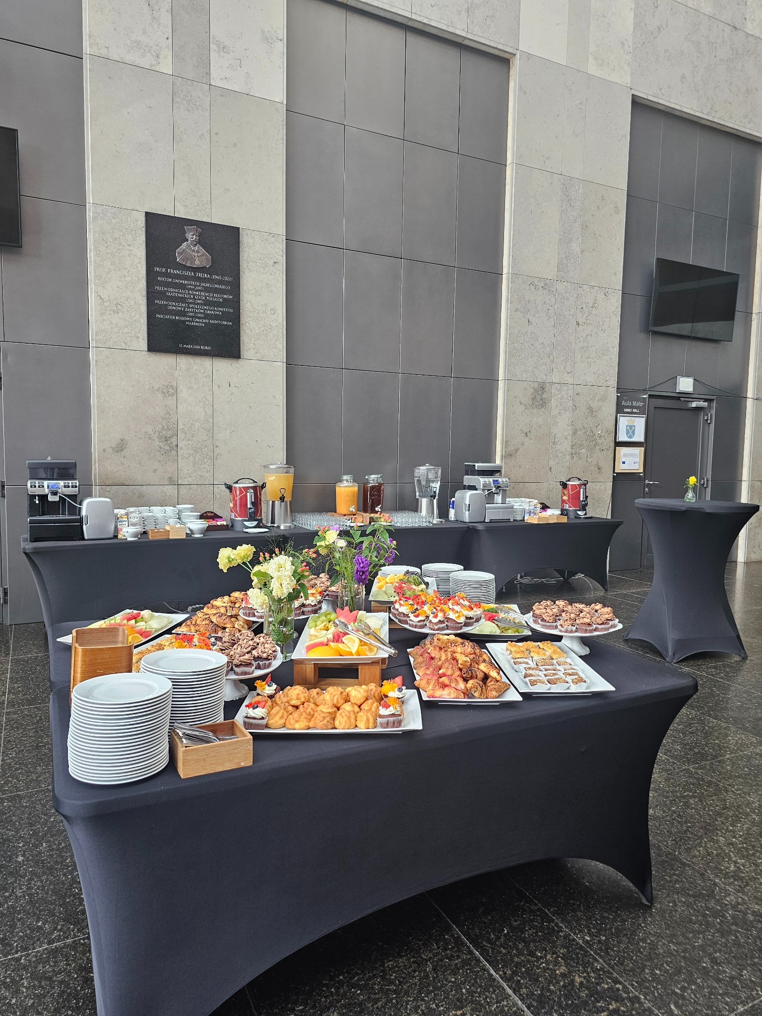 Stół z czarnym obrusem, zastawiony różnorodnymi przekąskami, ciastami i owocami na białych tacach, w tle ekspresy do kawy, soki i kwiaty, całość w nowoczesnym wnętrzu z szarą ścianą i tablicą...