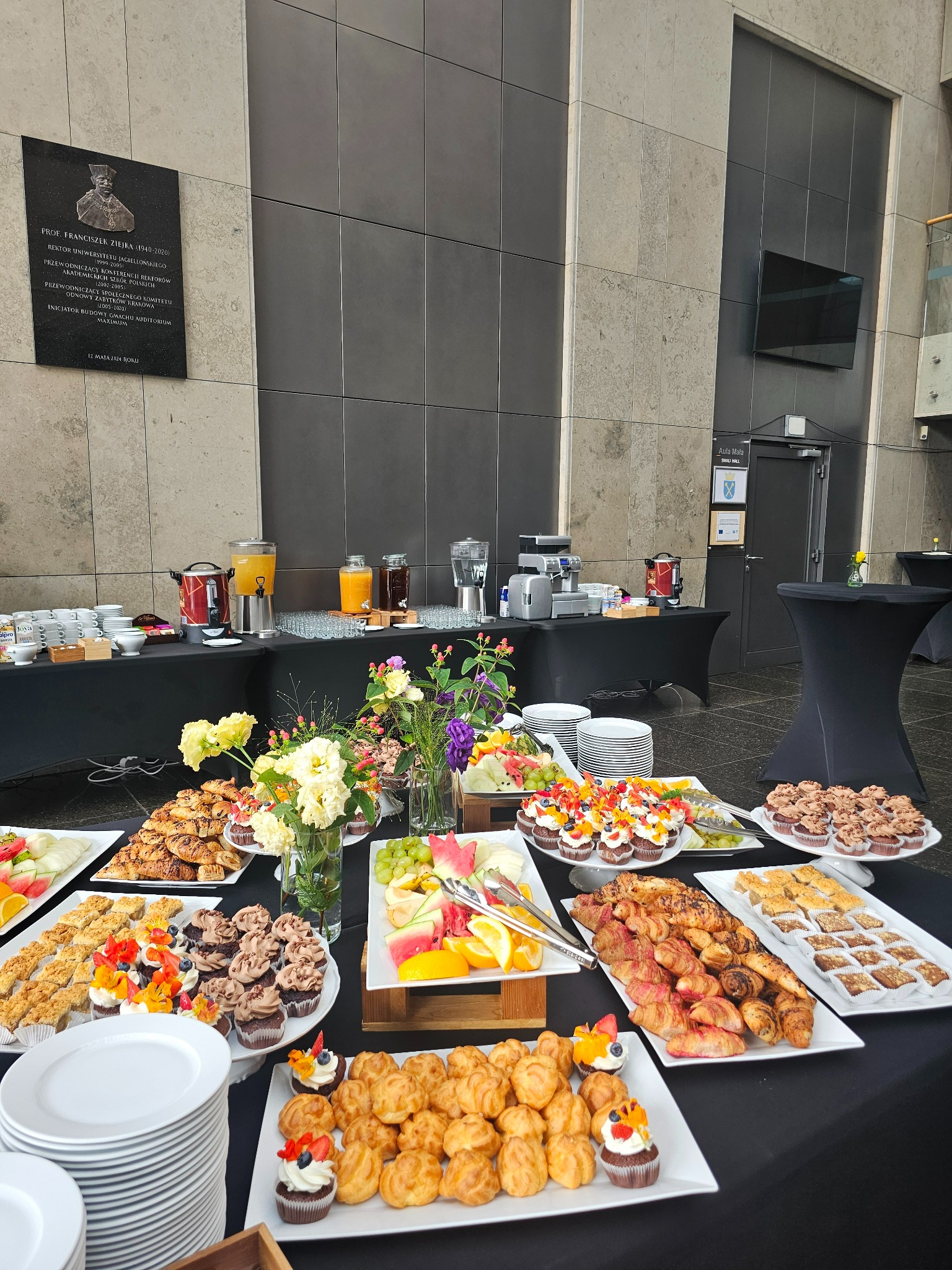 Stół z czarnym obrusem zastawiony różnorodnymi przekąskami: babeczki z owocami, ciastka francuskie, owoce pokrojone w plastry. W tle dystrybutory z sokami, kawa, kubki i talerze, na ścianie tablica...