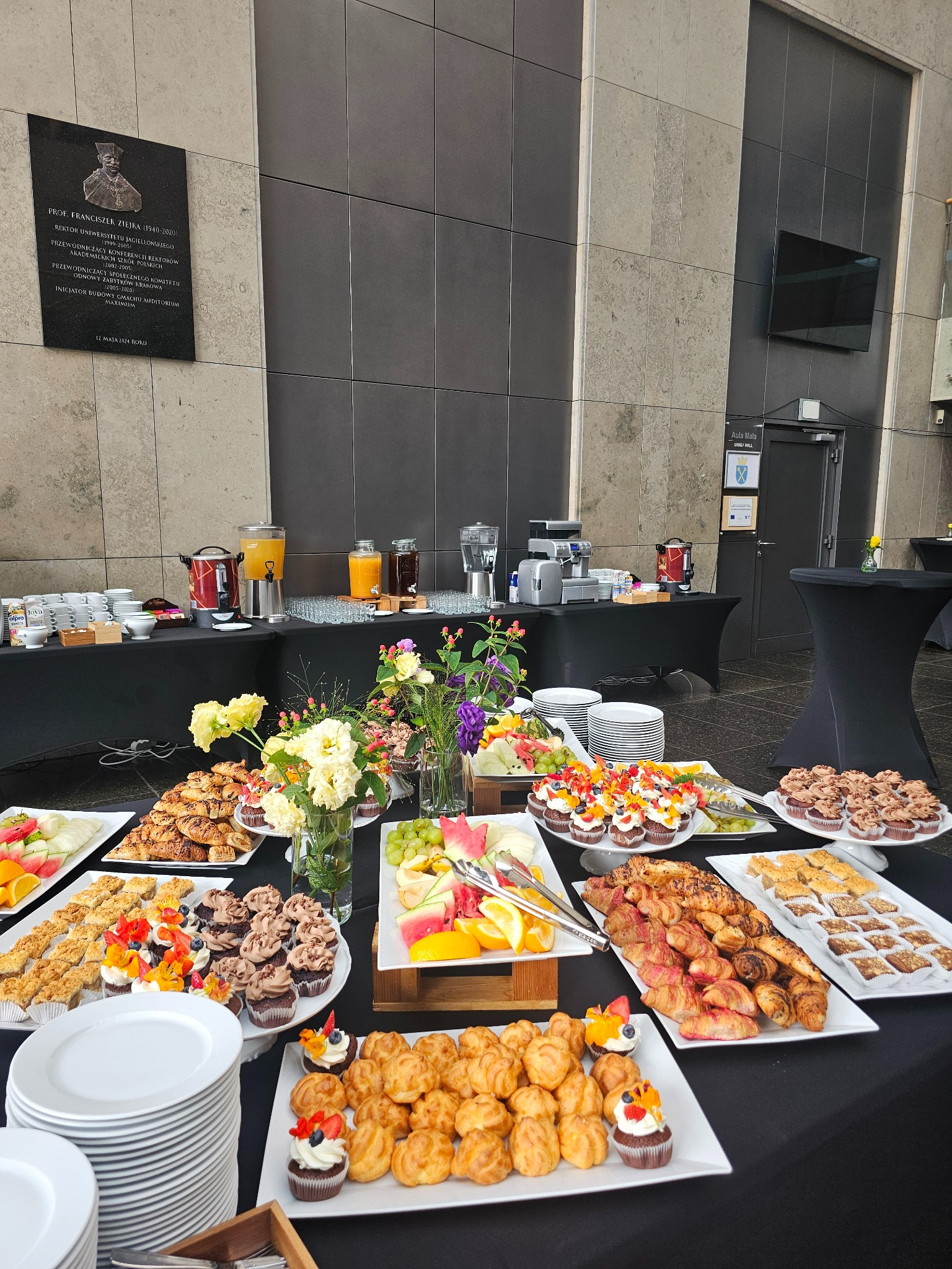 Stół cateringowy z różnorodnymi przekąskami, w tym babeczkami z owocami, ciastkami, rogalikami i owocami, ustawiony w przestronnym wnętrzu z kamienną ścianą i tablicą pamiątkową.