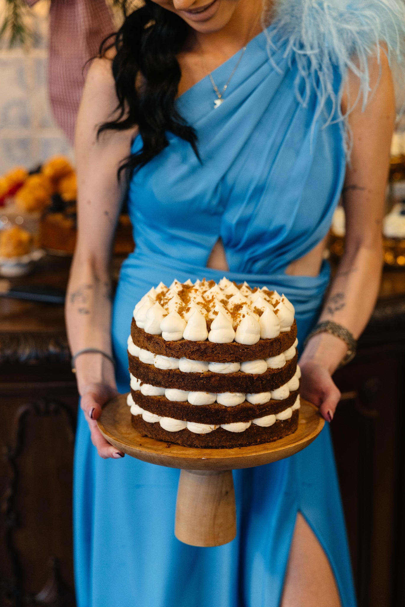 Kobieta w błękitnej sukni prezentuje tort z kremem na drewnianym stojaku, widoczne detale dekoracji i tło z elementami zastawy.