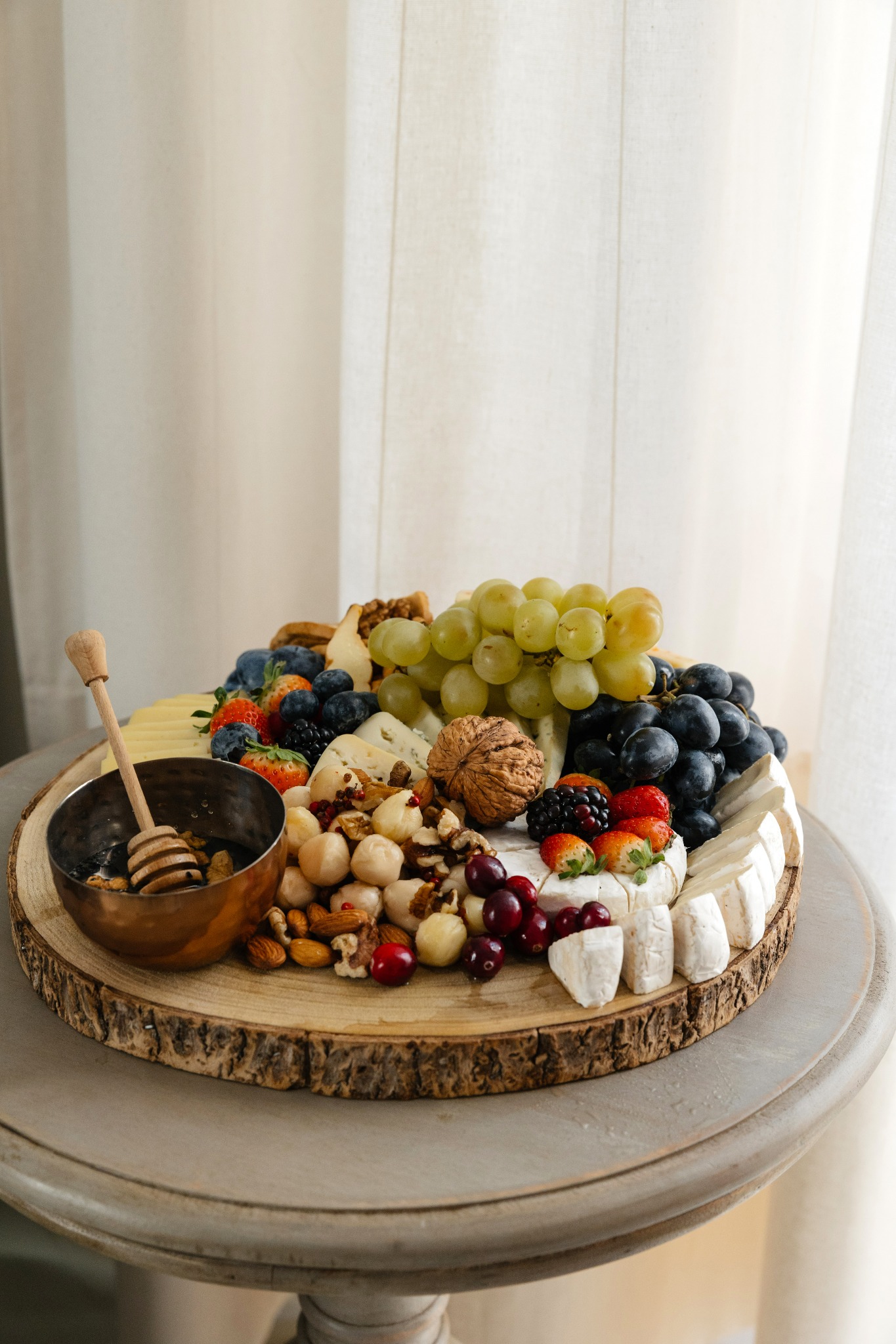Elegancka deska serów, owoców i orzechów na drewnianym talerzu w Krakowie, z miodem w miedzianej miseczce i drewnianą łyżeczką, na tle kremowej zasłony.
