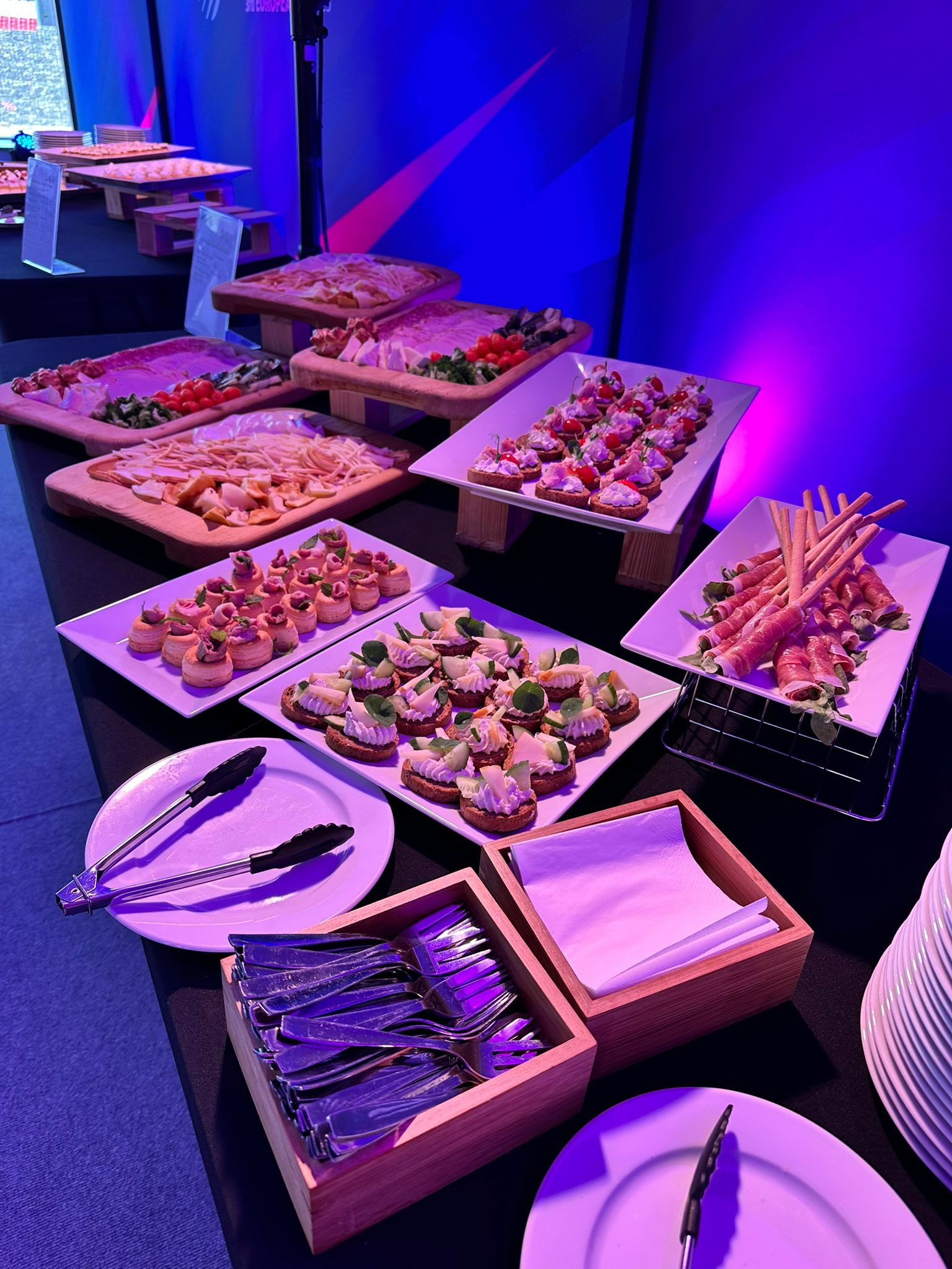 Stół z przekąskami na przyjęciu: kanapeczki z różnymi dodatkami, grissini owinięte szynką, deski serów i wędlin, sztućce i talerze, oświetlenie w odcieniach fioletu.