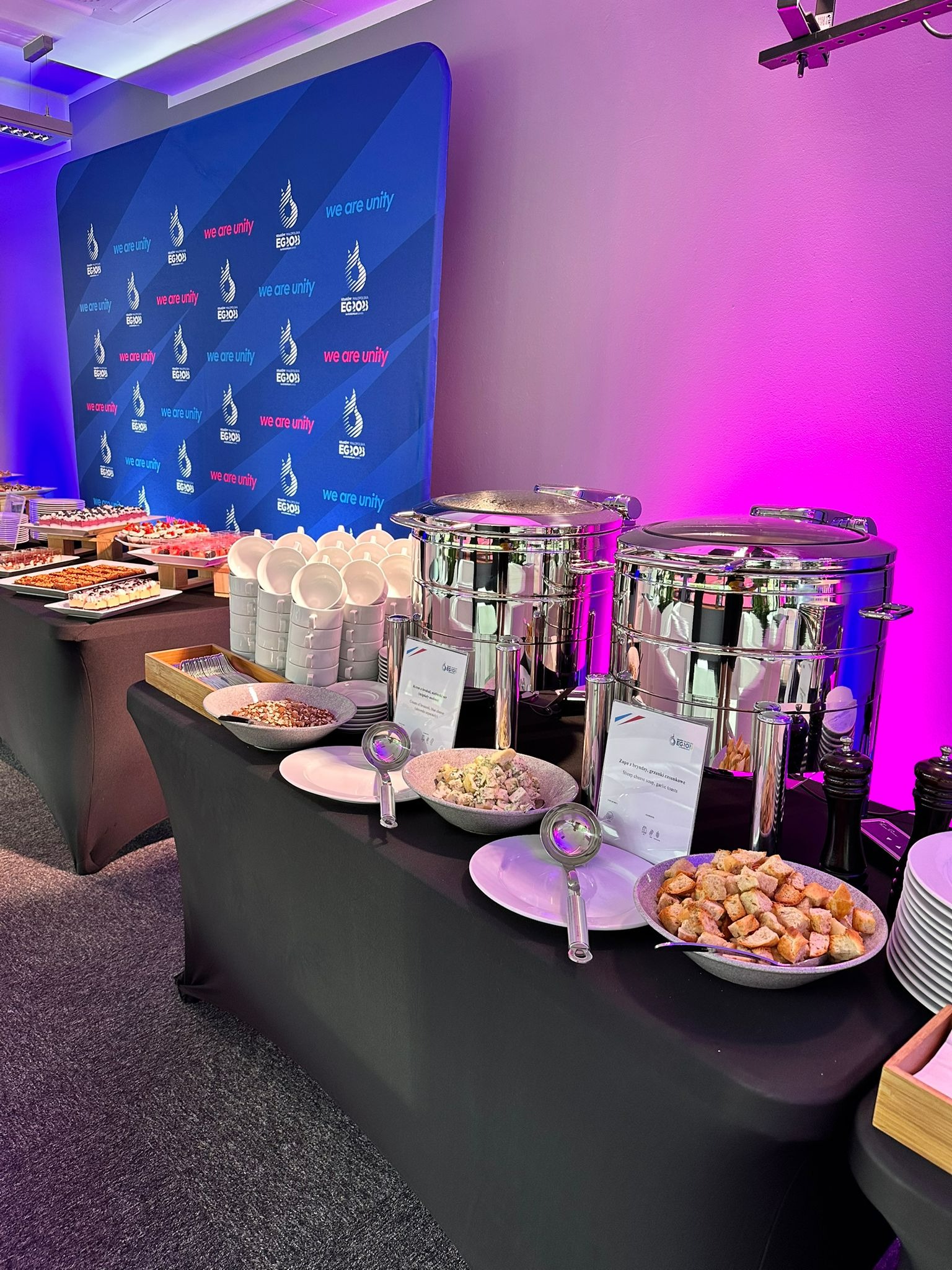 Stół cateringowy z metalowymi bemarami, zupą z grzankami czosnkowymi, sałatkami i deserami, tło z logo firmy podświetlone fioletowym światłem.