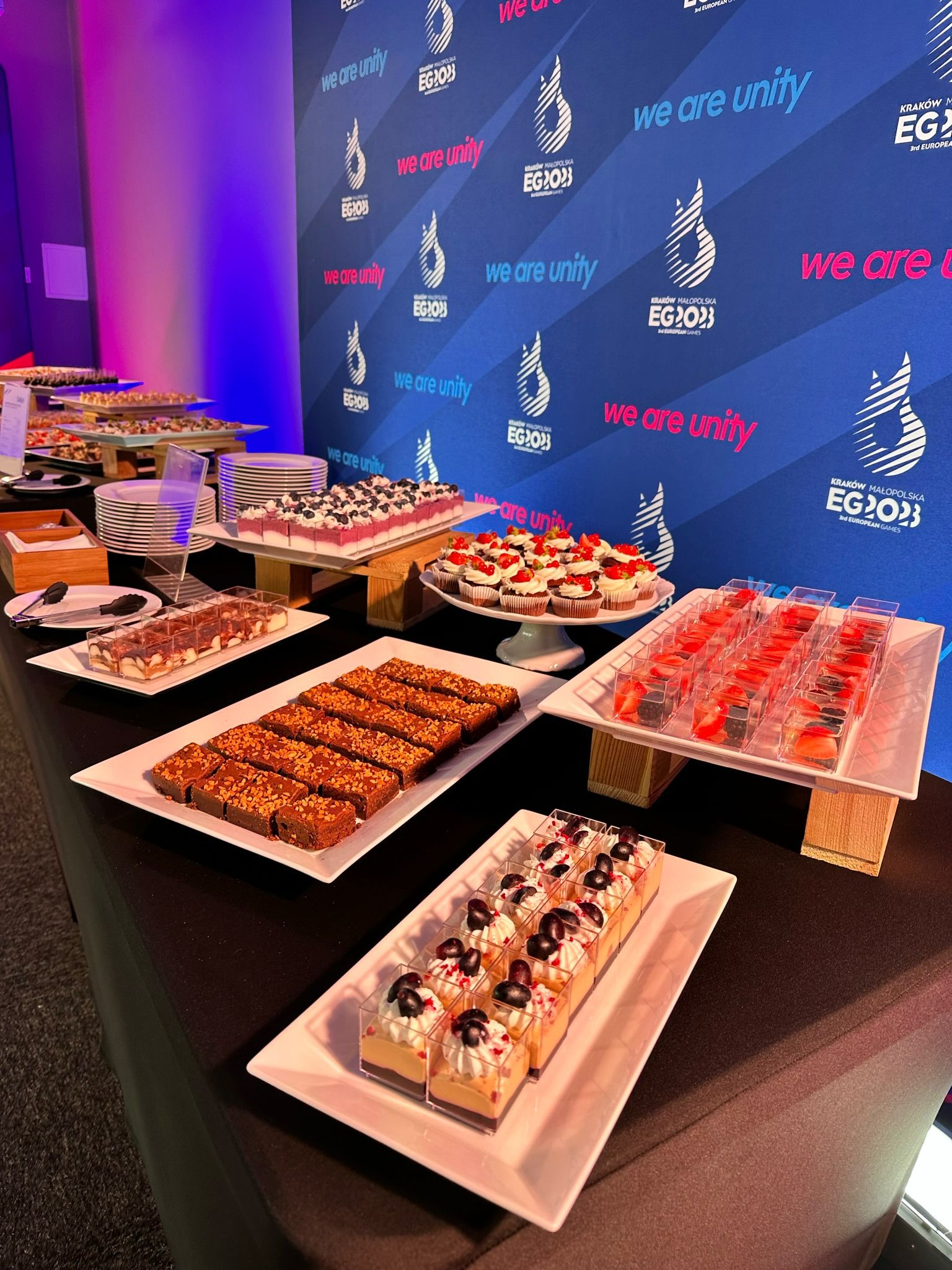 Stół z różnorodnymi deserami, w tym brownie z orzechami, mini serniki z borówkami, babeczki z truskawkami i desery w przezroczystych pojemnikach z owocami, ustawiony na tle z logo 'Kraków...