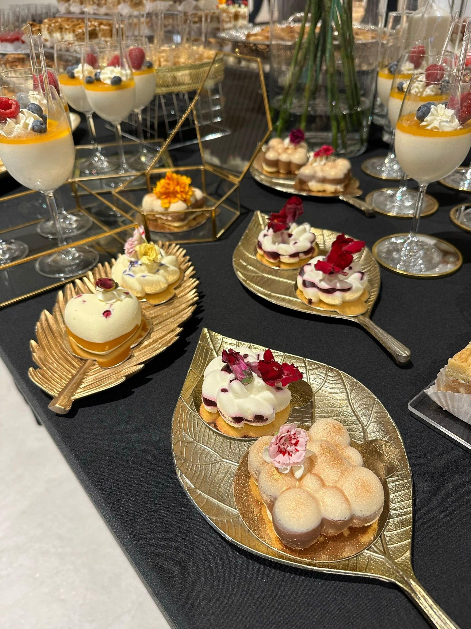 Elegancko zaaranżowany stół z deserami: panna cotta z owocami w szklankach, mini torty na złotych podstawkach w kształcie liści, dekorowane kwiatami.