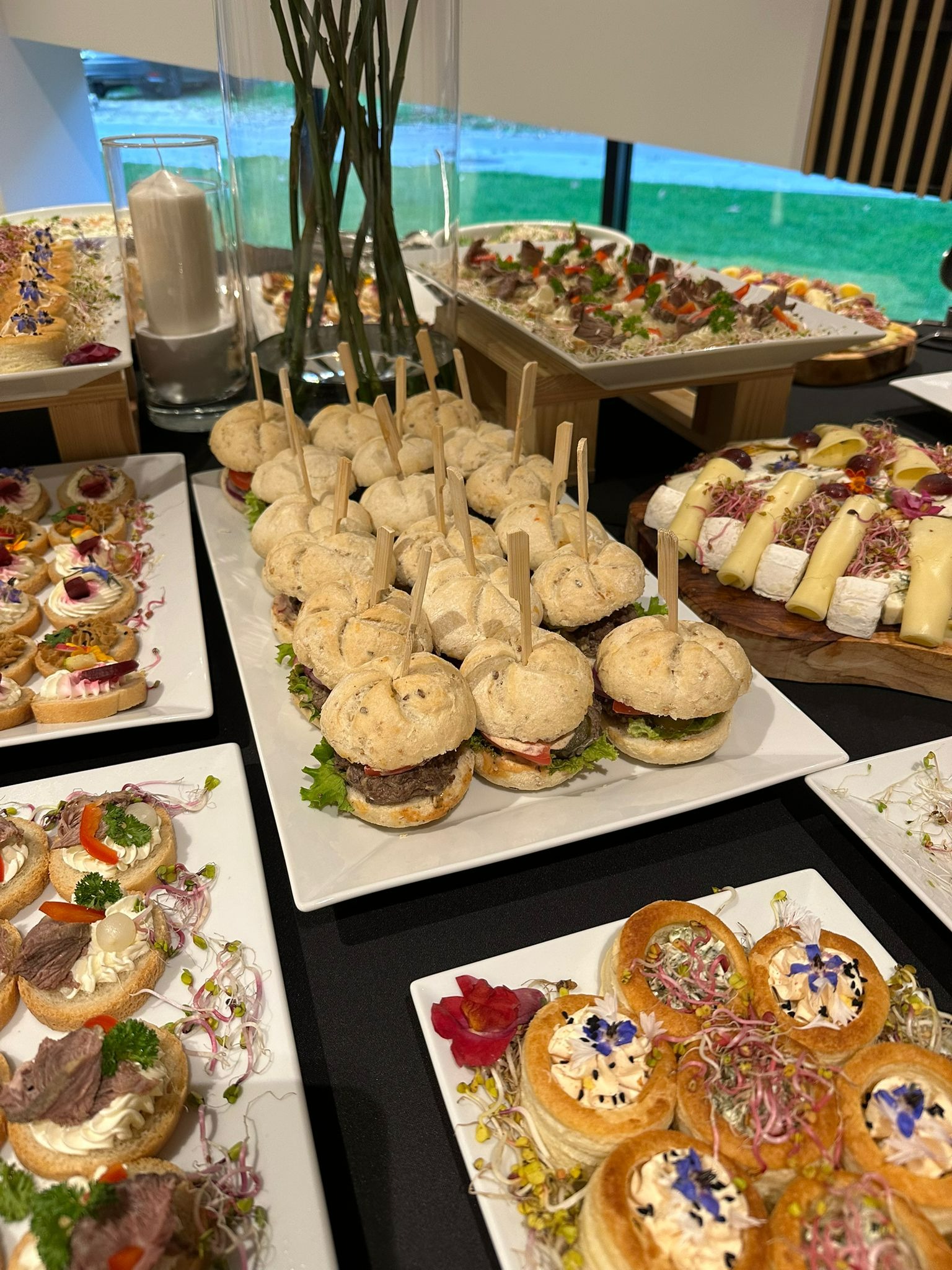 Stół z różnorodnymi przekąskami: mini burgery z wykałaczkami, tartaletki z kremem i kwiatami, kanapeczki z mięsem, deska serów i sałatka na tle okna.