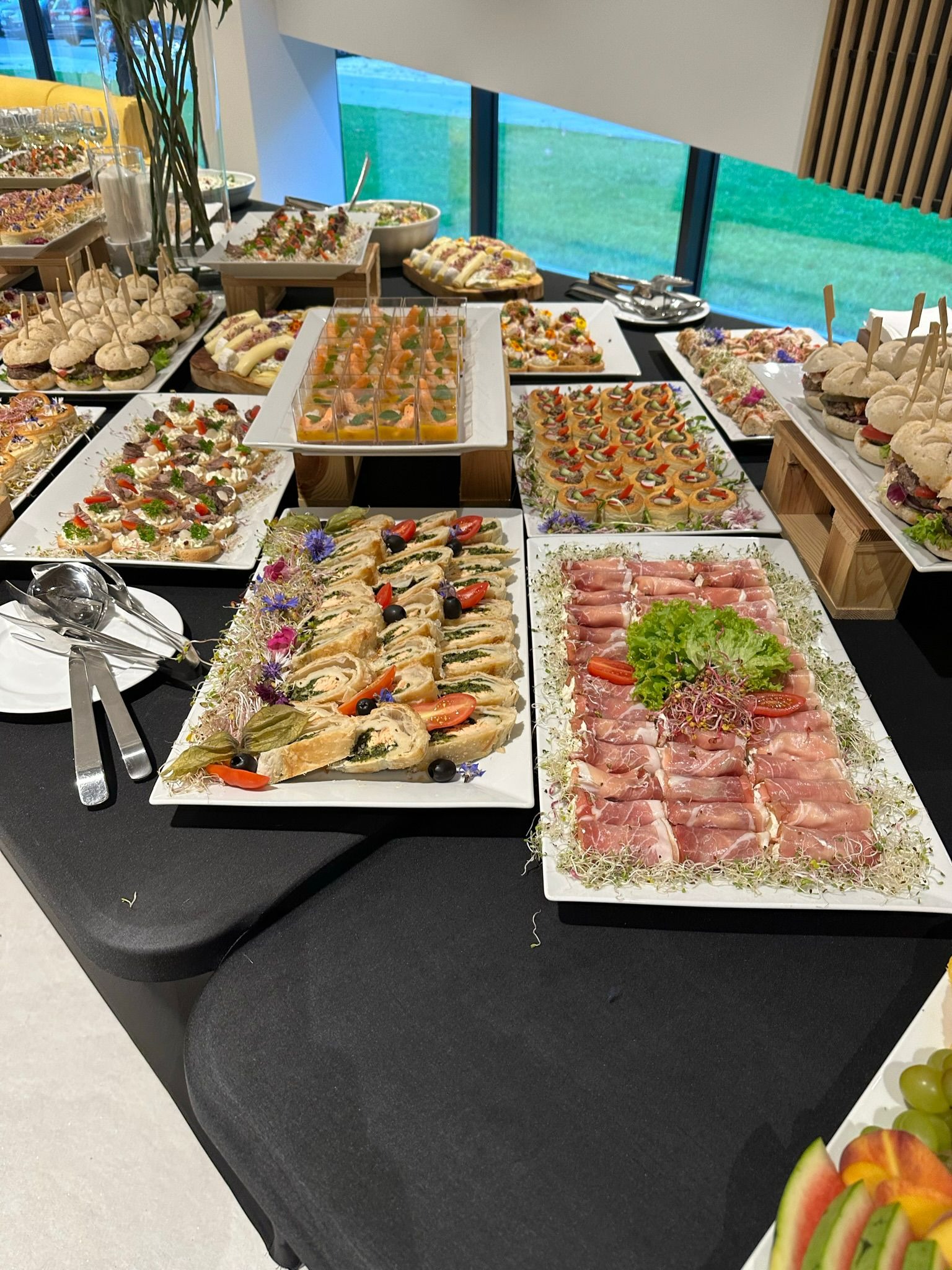 Stół z czarnym obrusem, na którym znajdują się różnorodne przekąski na białych talerzach, w tym roladki z nadzieniem, szynka parmeńska, mini burgery i tartaletki udekorowane kwiatami i kiełkami.