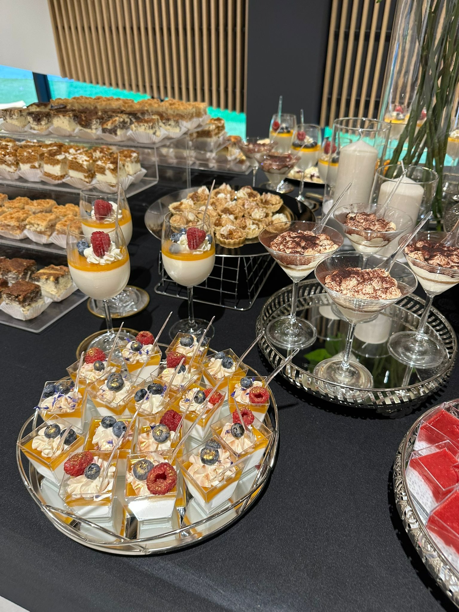 Bogato zastawiony stół z deserami: tiramisu w kieliszkach, galaretki z owocami, ciasta kremowe i panna cotta z malinami i borówkami, ułożone na srebrnych tacach na czarnym obrusie.