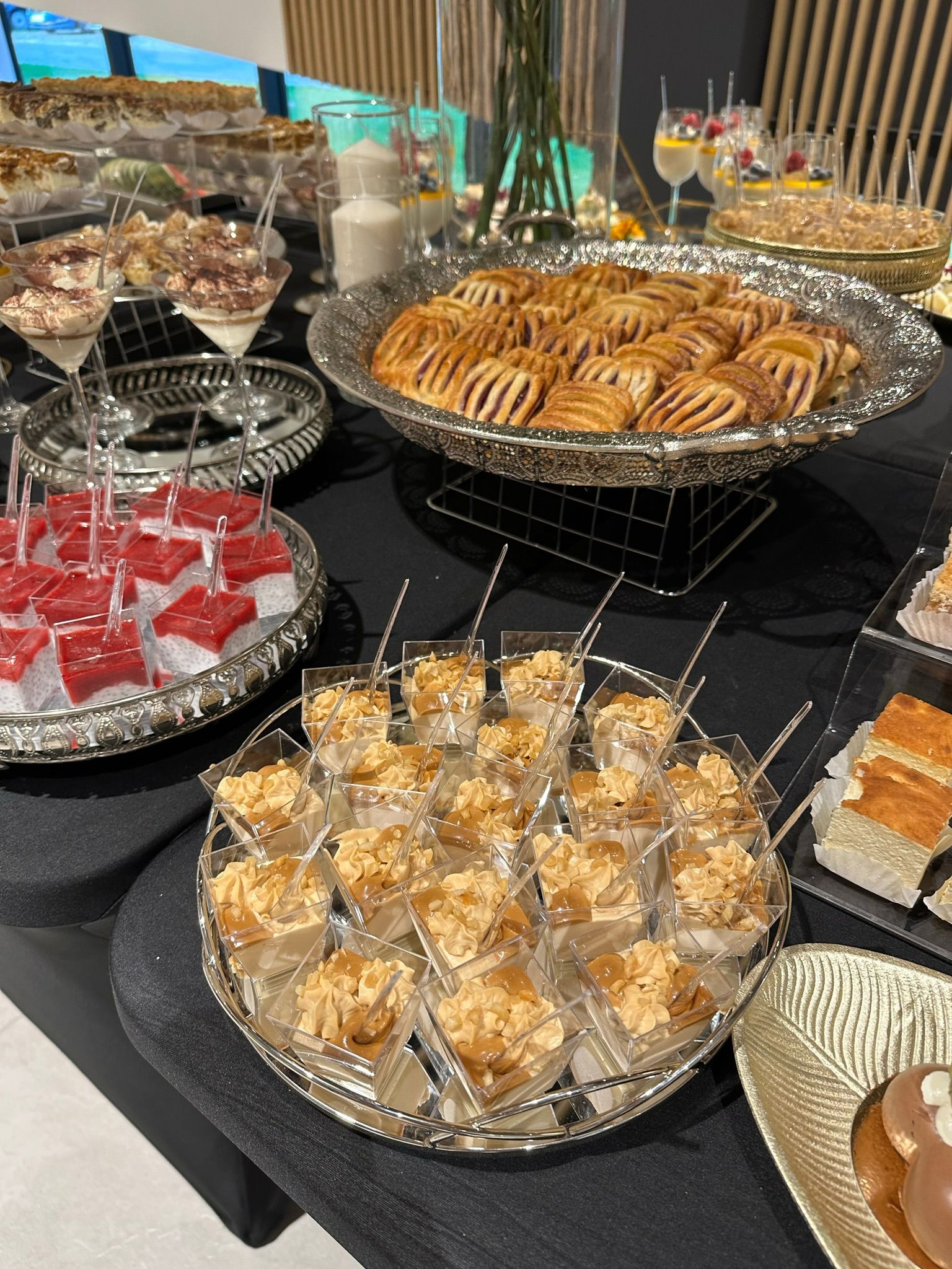 Elegancki bufet słodkości z różnorodnymi deserami w szklanych naczyniach i srebrnych paterach, na czarnym obrusie, z widocznymi tartaletkami z dżemem, deserami z musem i owocami oraz ciastami.