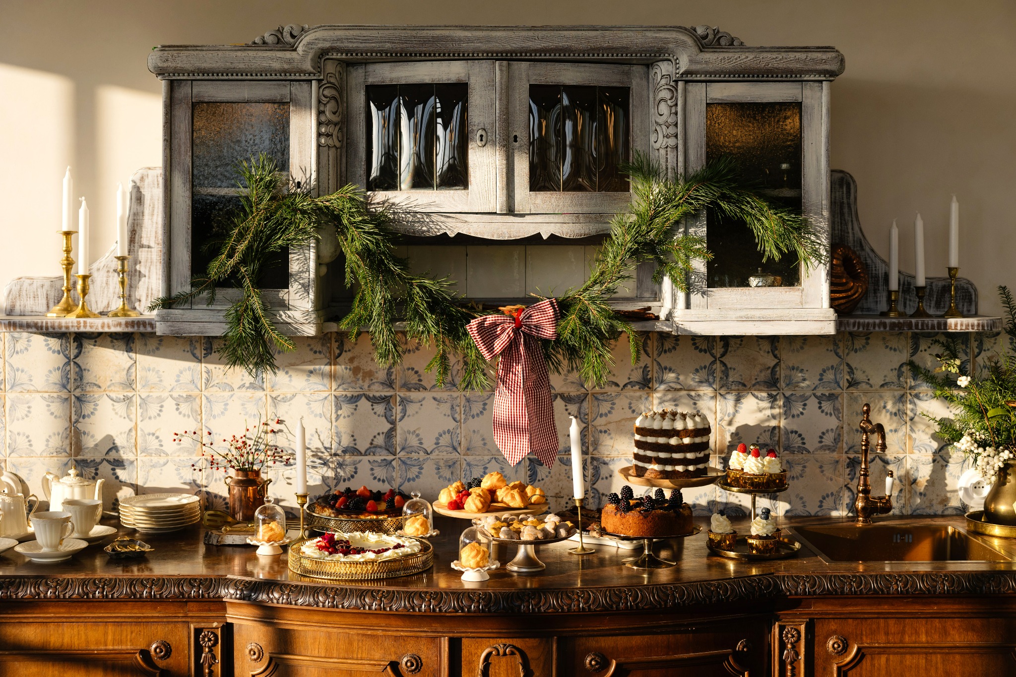 Świąteczny stół z ciastami, tortami i deserami w rustykalnej aranżacji, ozdobiony gałązkami świerku i czerwoną kokardą w kratę, na tle szafki z witrynami i ceramicznych płytek.
