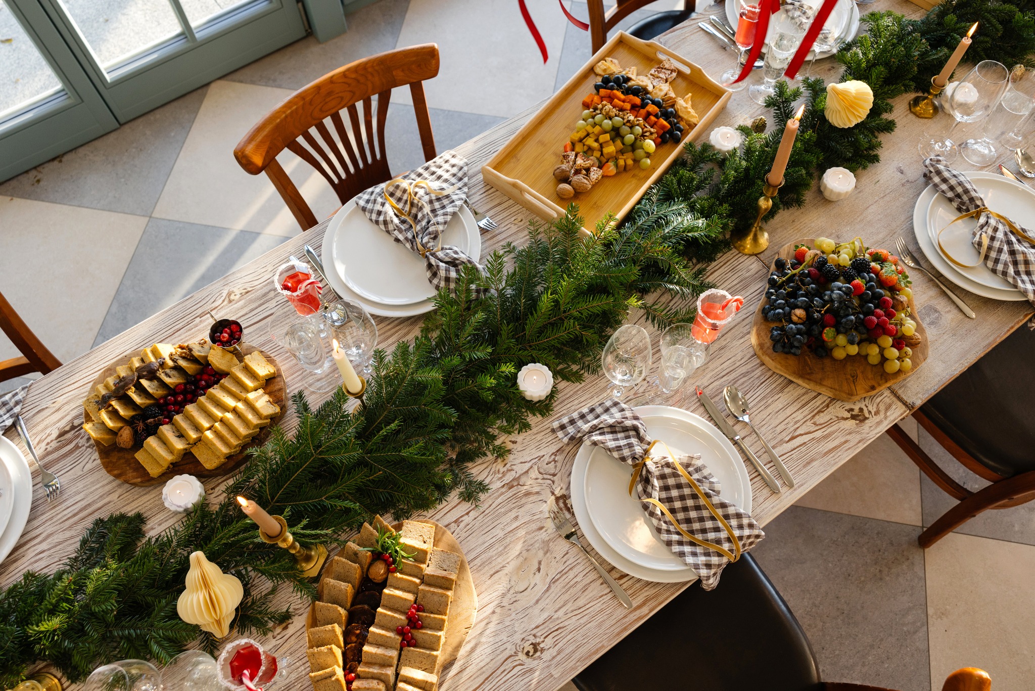 Uroczysty stół z drewnianym blatem, udekorowany świerkowymi gałązkami, świecami w złotych świecznikach, talerzami z kostkowanymi serwetkami i różnorodnymi przekąskami, w tym ciastami, owocami...