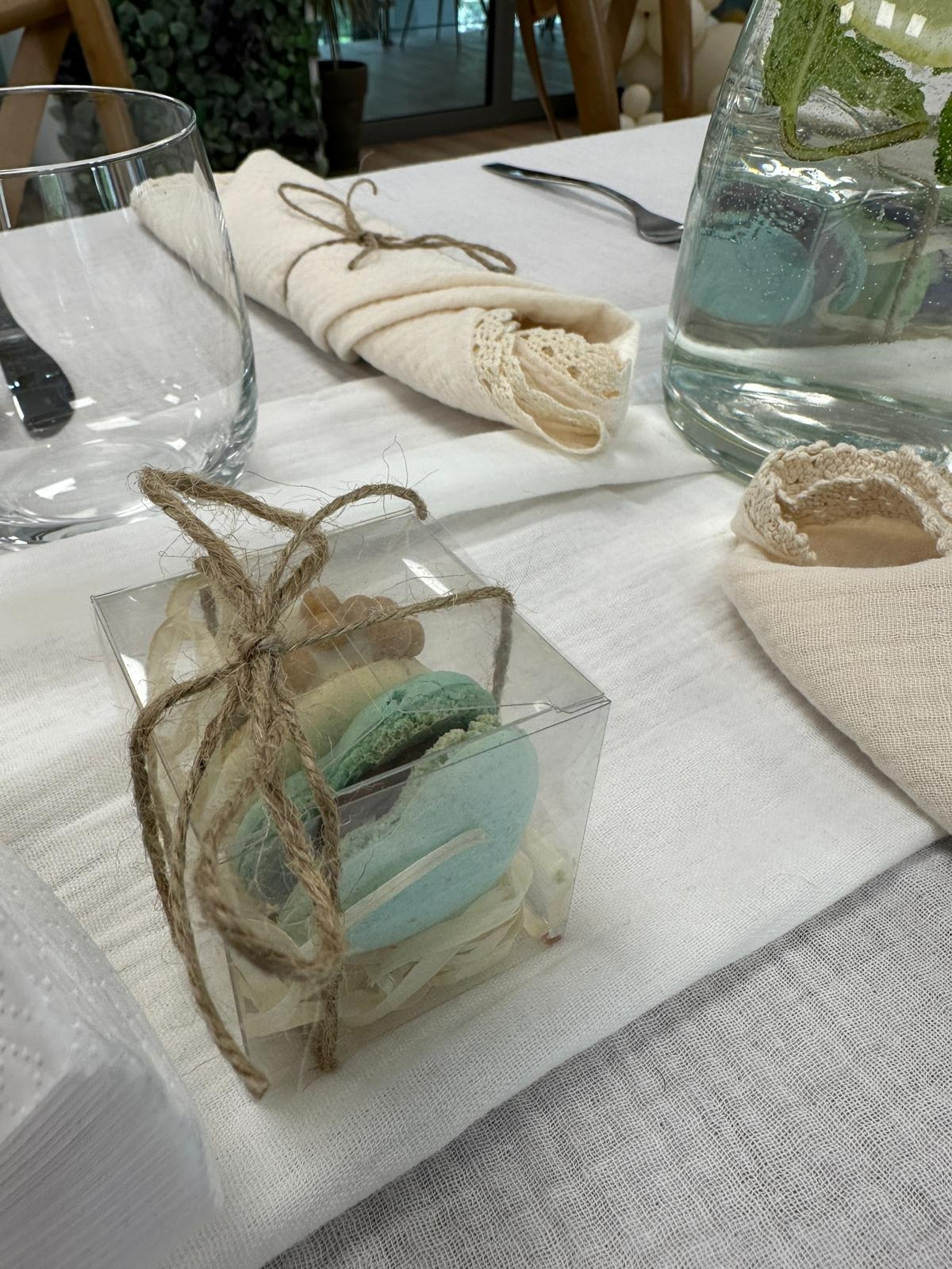 Ustawienie stołu z dekoracyjnymi serwetkami z koronką, szklanką, dzbankiem z wodą i pudełeczkiem z makaronikami związanym sznurkiem.