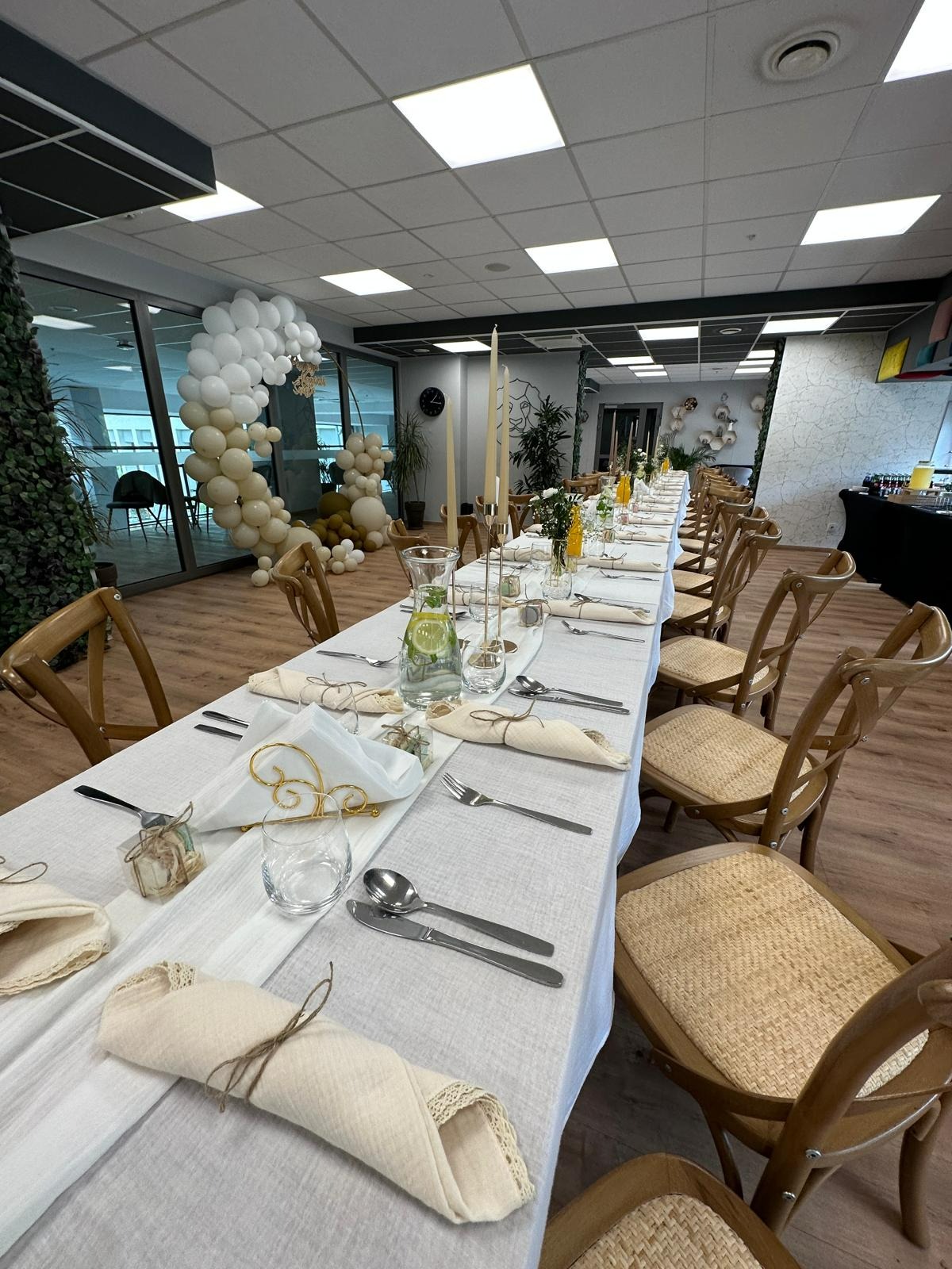 Uroczysty stół nakryty białym obrusem, z beżowymi serwetkami związanymi sznurkiem, srebrnymi sztućcami, szklanymi naczyniami, świecami w złotych świecznikach, dekoracjami kwiatowymi i balonową...