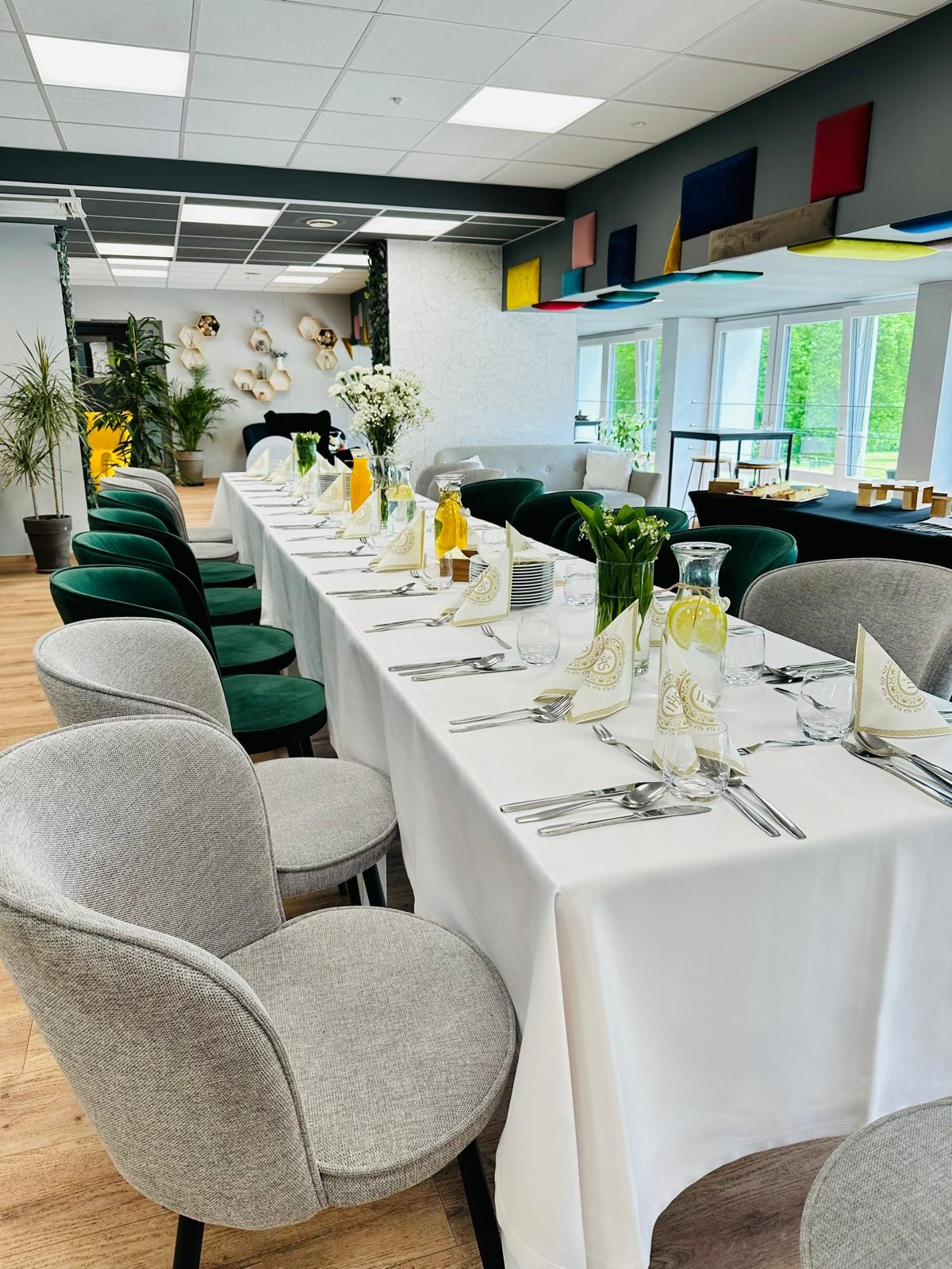 Długi stół nakryty białym obrusem, z elegancką zastawą, szklanymi karafkami z cytryną i kwiatami, otoczony krzesłami w odcieniach szarości i butelkowej zieleni, w jasnym wnętrzu z dekoracjami...