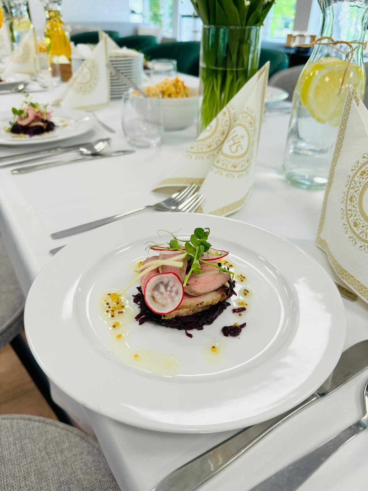 Elegancko podane danie na białym talerzu: pieczone mięso na purpurowej kapuście, udekorowane plasterkiem rzodkiewki, jabłkiem i mikroziołami, polane sosem z gorczycą, na stole z białym obrusem...