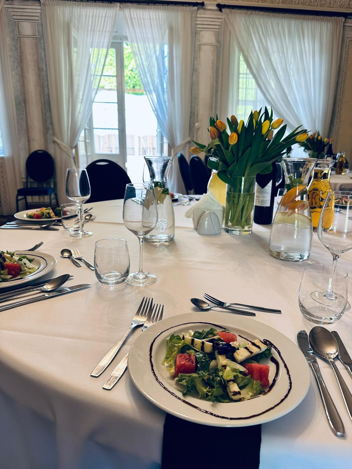 Elegancko nakryty stół z sałatkami, napojami i bukietami żółtych tulipanów w jasnym pomieszczeniu z oknami.