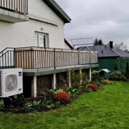 Biała jednostka zewnętrzna klimatyzatora Haier zamontowana przy schodach tarasu domu jednorodzinnego, widok na zadbany ogród z trawnikiem i dekoracyjnymi rabatami.