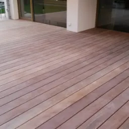 Nowoczesny taras z desek w odcieniach brązu, ułożonych równolegle do przeszklonych drzwi balkonowych, z widocznym fragmentem ogrodu i białym oświetleniem kulistym w tle.