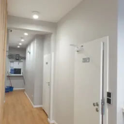 Korytarz biurowy z drewnianą podłogą, białymi ścianami i drzwiami z napisem 'Pomieszczenie socjalne', oświetlony lampami sufitowymi