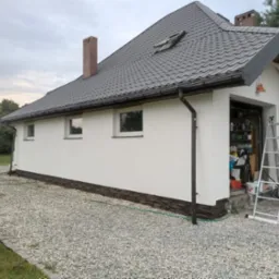 Świeżo pomalowana biała elewacja domu jednorodzinnego z szarą dachówką, widoczne okna, rynny, fragment garażu z drabiną i stosem opon na żwirowym podłożu.