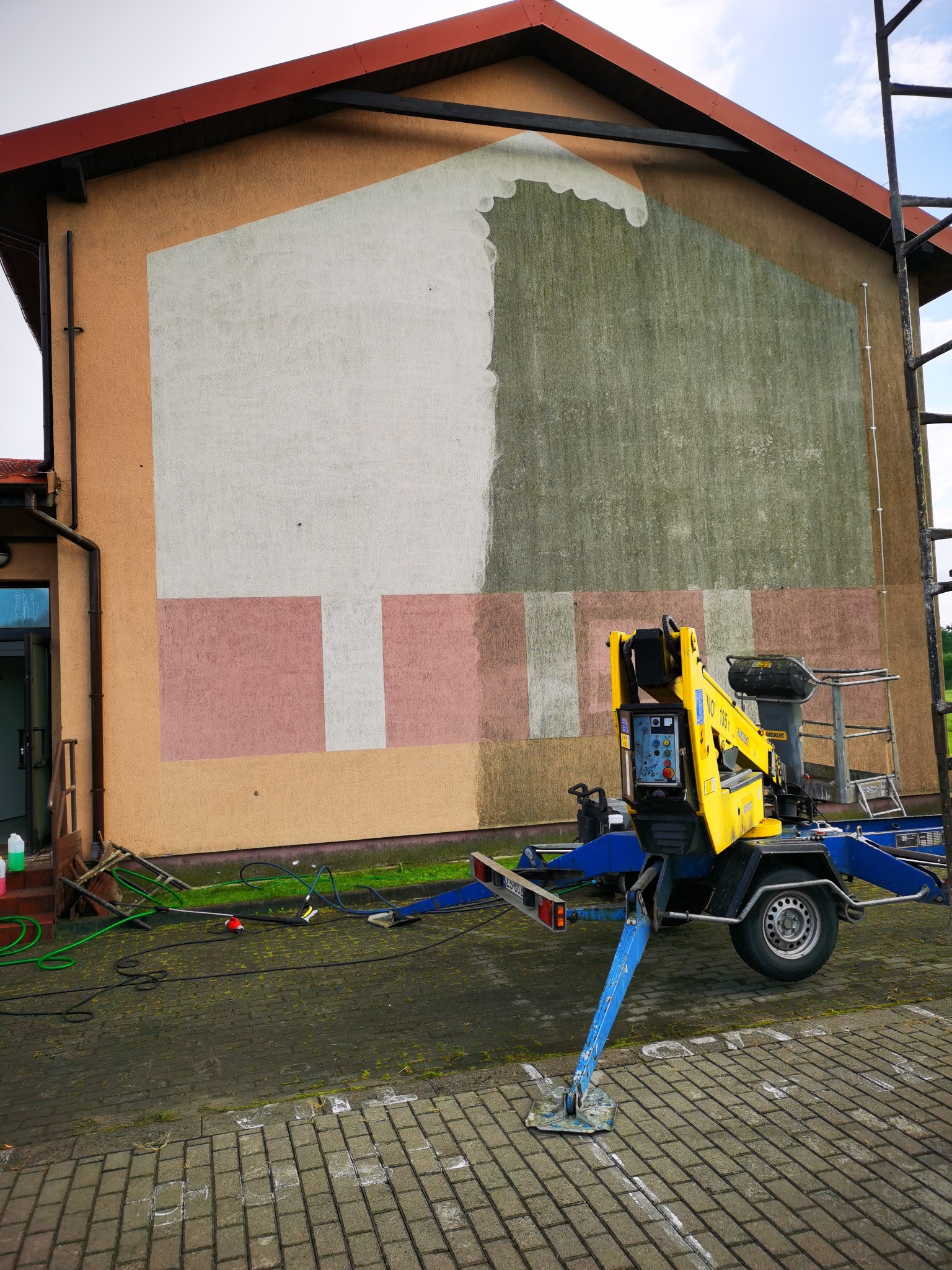 Żółty podnośnik koszowy na tle częściowo umytej, wielokolorowej elewacji budynku. Widoczna różnica w kolorze i czystości pomiędzy umytą a nieumytą częścią ściany.