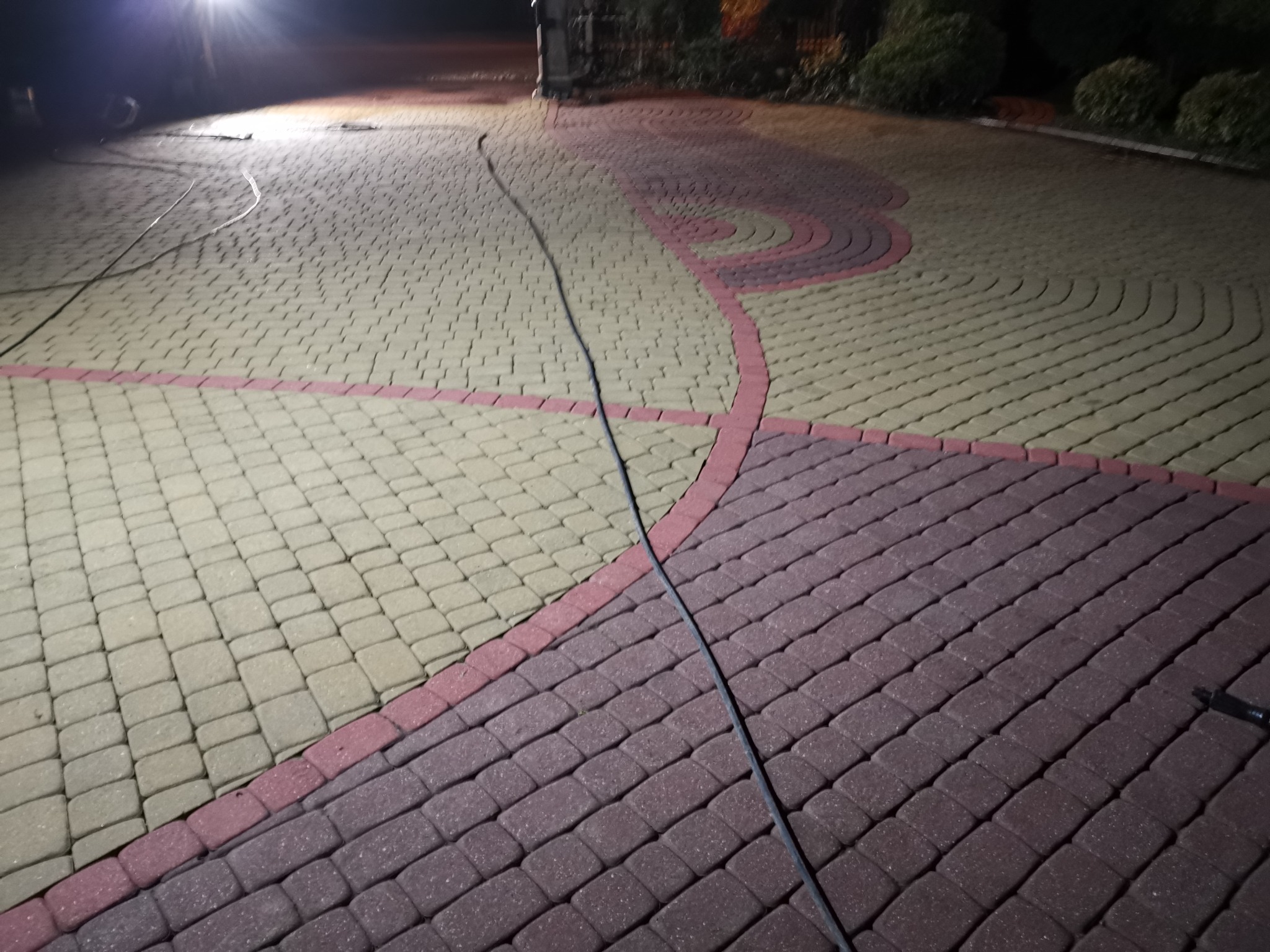 Ułożona kostka brukowa w dwóch kolorach z dekoracyjnym wzorem, oświetlona reflektorem w nocy, z widocznym kablem zasilającym.